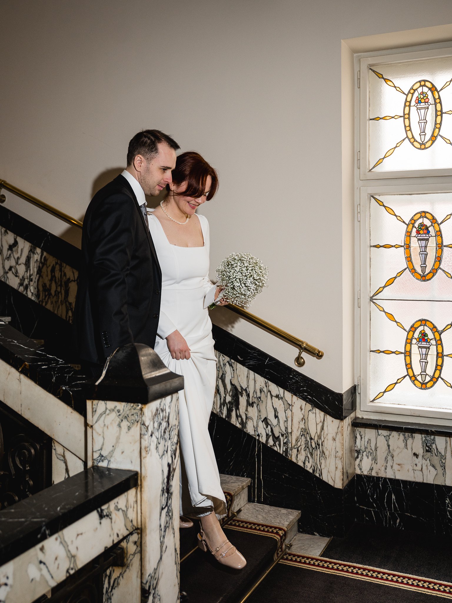 A bride and groom descending a staircase indoors, with stained glass windows nearby. The bride is wearing a white wedding dress and holding a bouquet, while the groom is in a black suit.