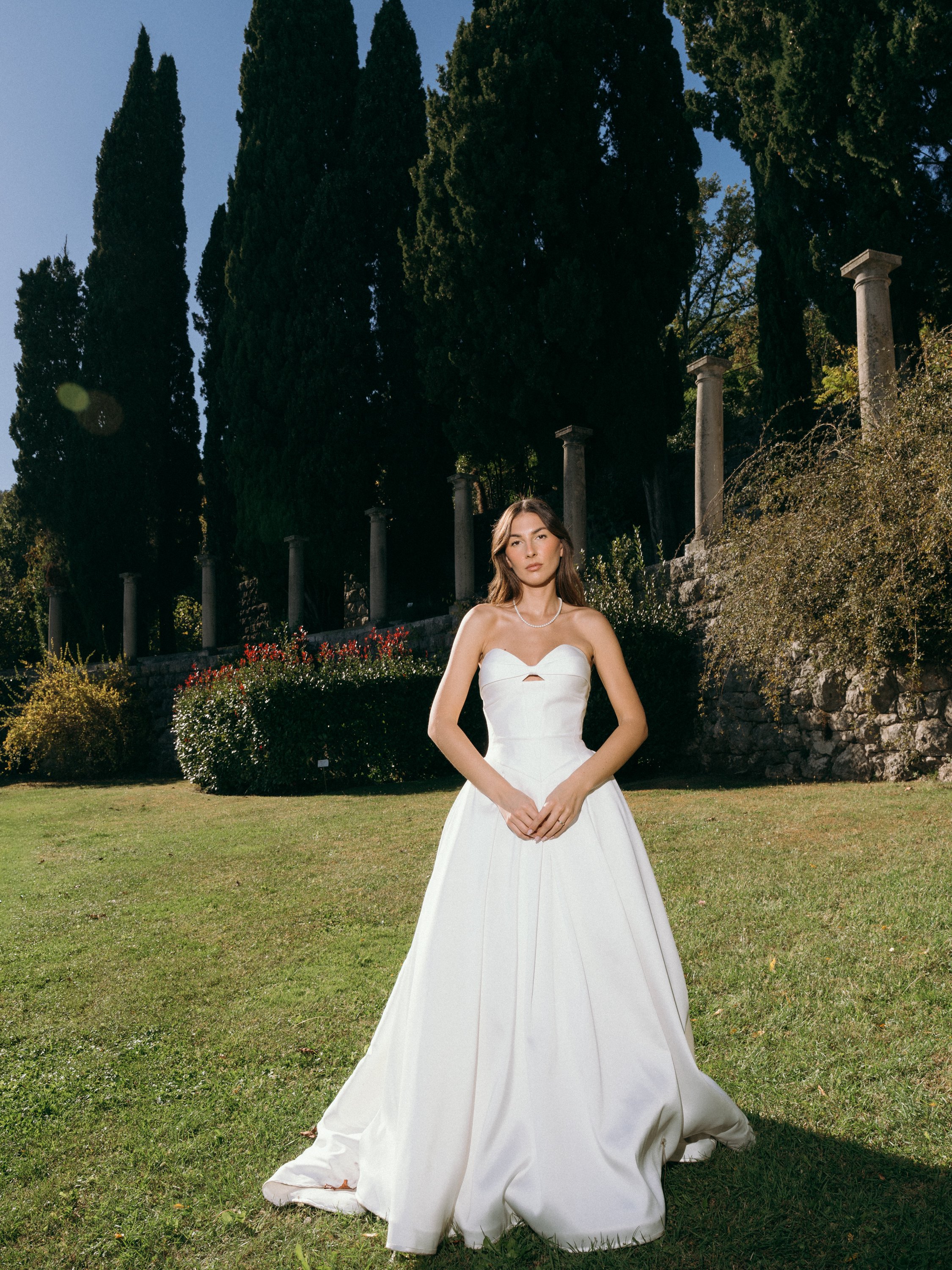 A woman in a white strapless gown standing on grass in a garden with tall trees and stone columns in the background.