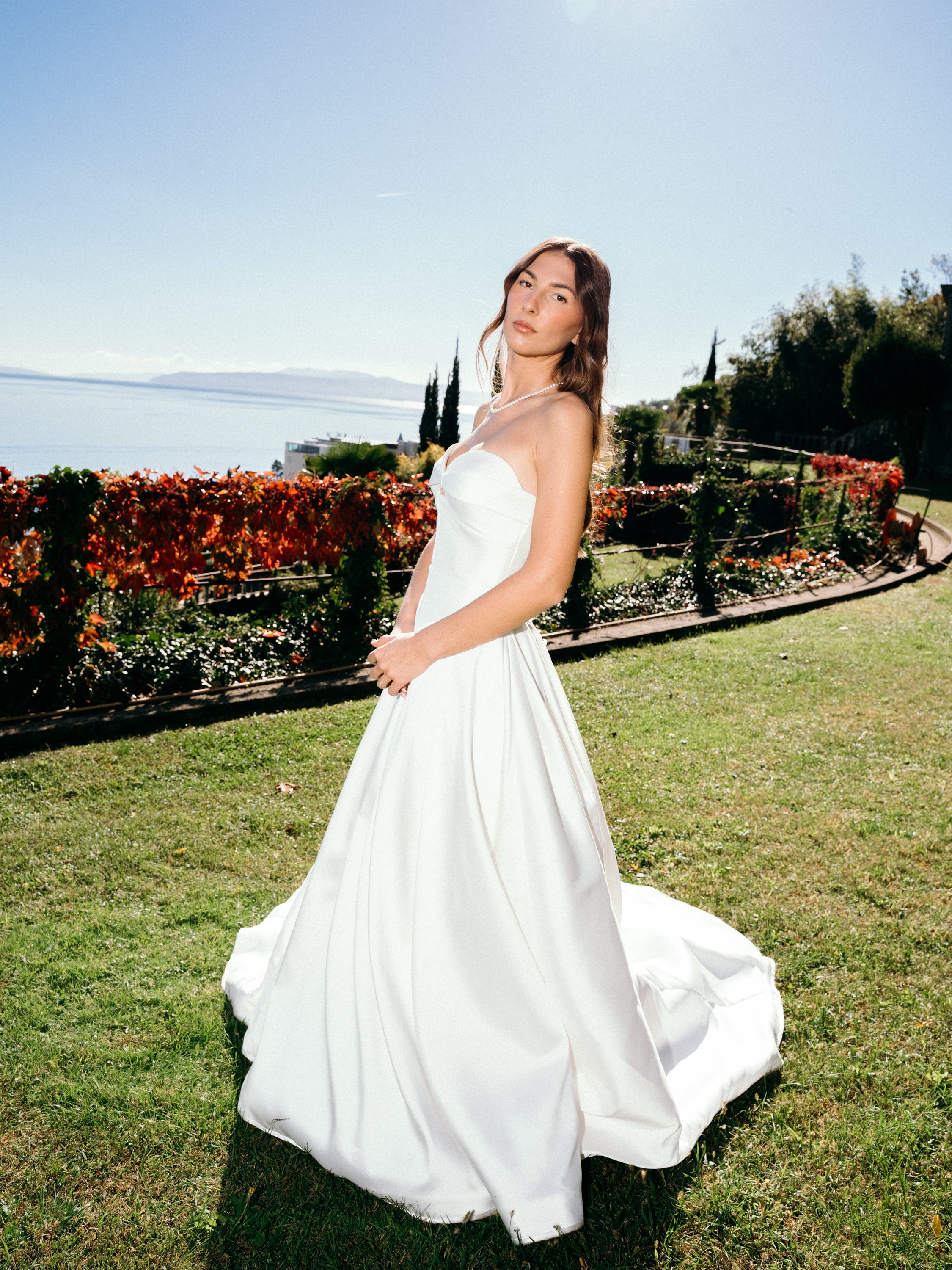 A woman in a strapless white wedding dress standing on a grassy area outdoors with a scenic view of water and distant land in the background.