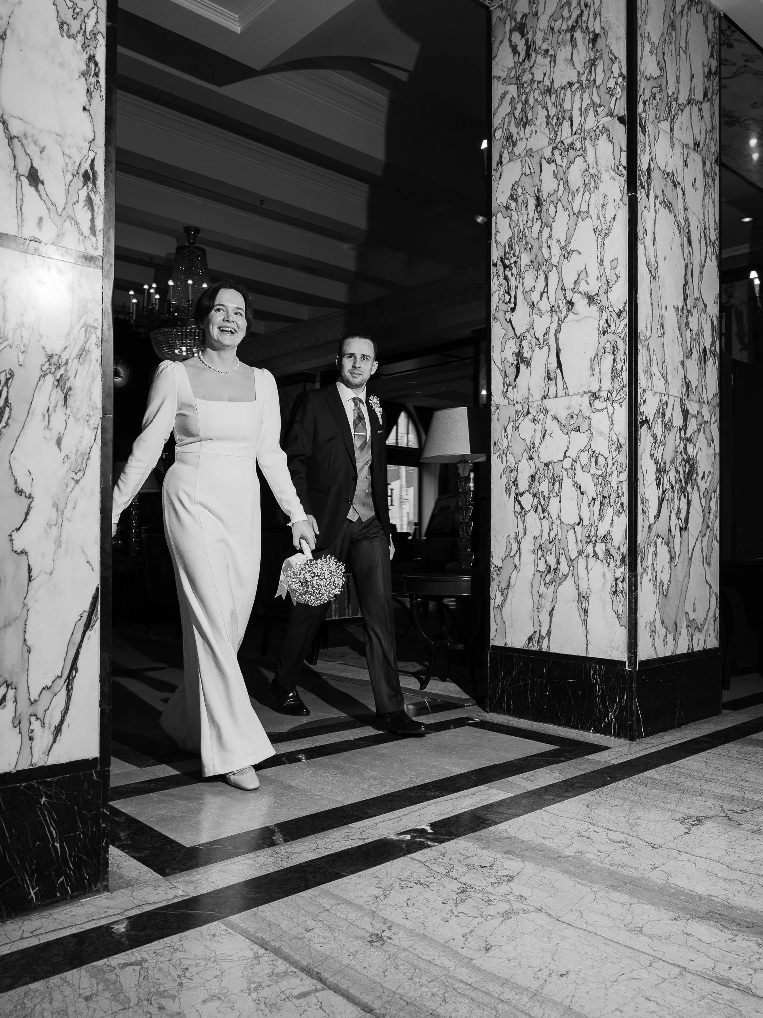 A woman and a man, dressed in wedding attire, walking through a marble doorway in an elegant building, possibly a hotel or event venue.