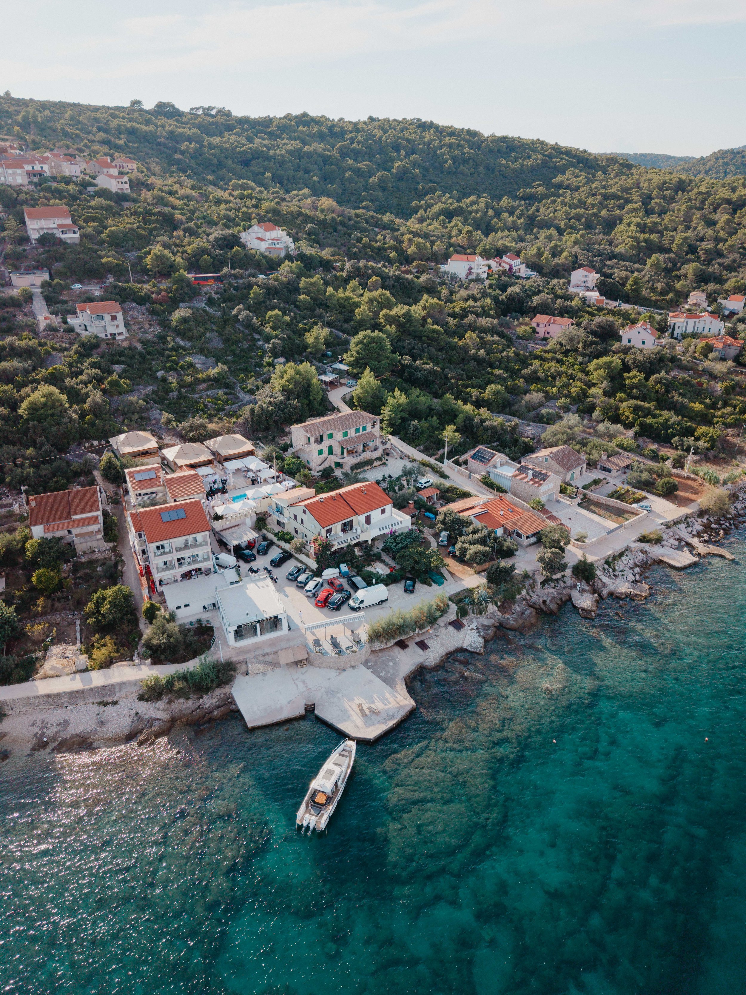 Aerial view of a hillside neighborhood near the sea with houses, trees, and a dock with a boat.