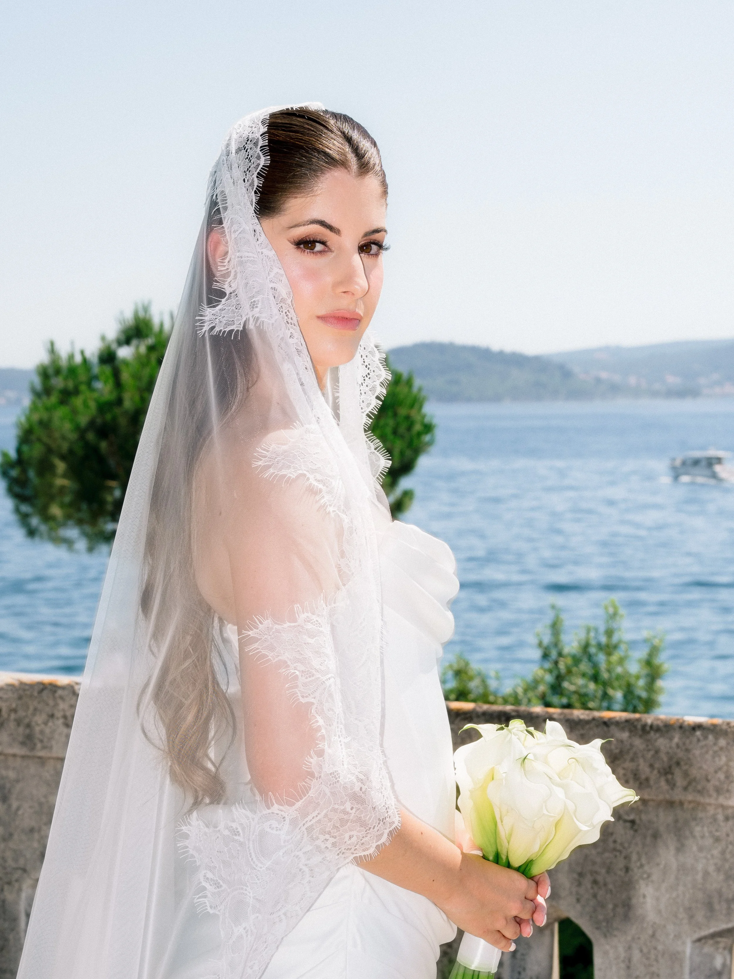 A bride in a white wedding dress and lace veil holding a bouquet of white flowers, standing outdoors near a body of water with hills in the background.