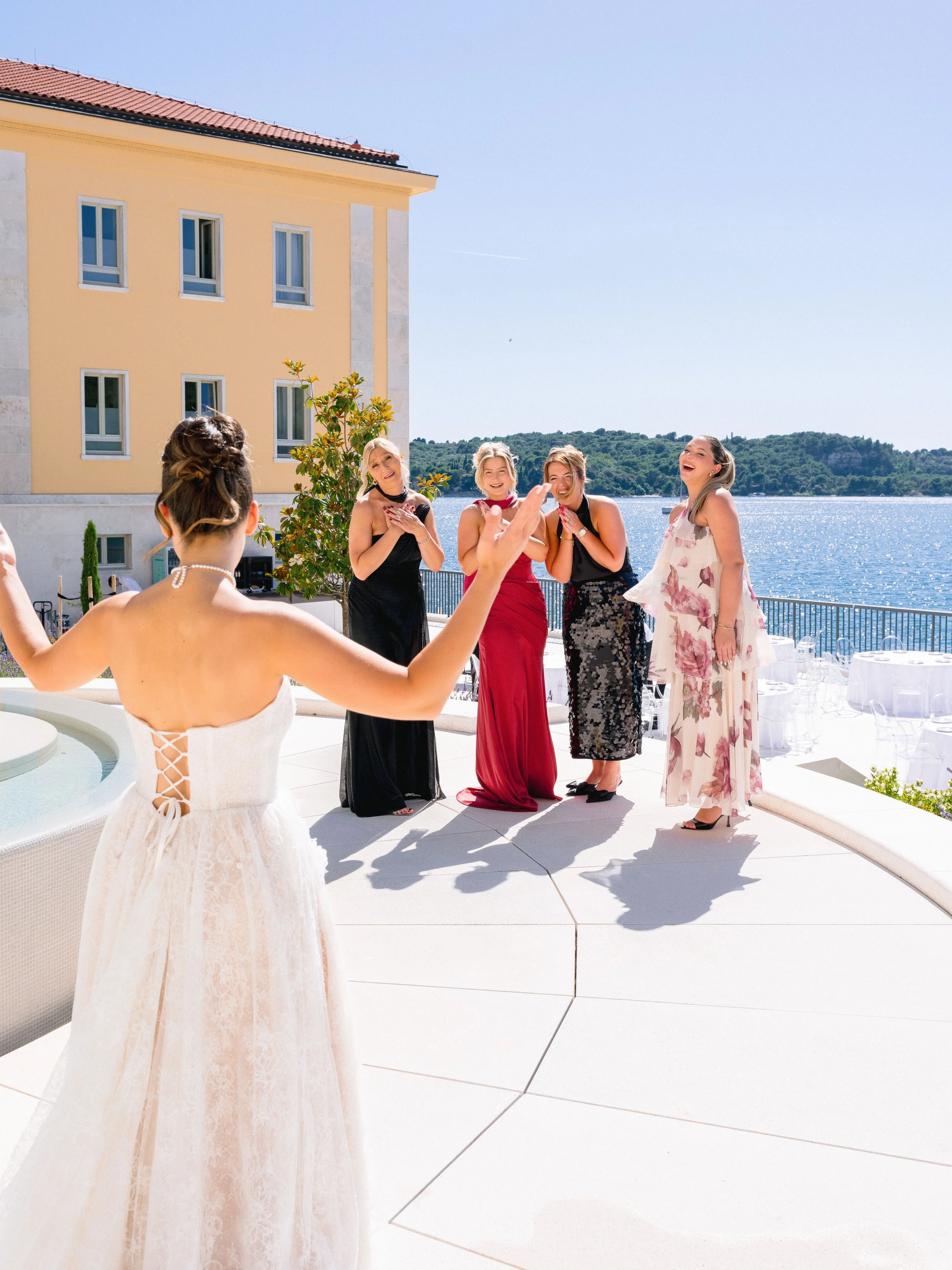 A bride in a white dress standing with her back to the camera, facing a group of five women who are laughing and smiling outdoors near a body of water. The women are dressed in evening gowns of various colors, and the scene appears to be at a celebra