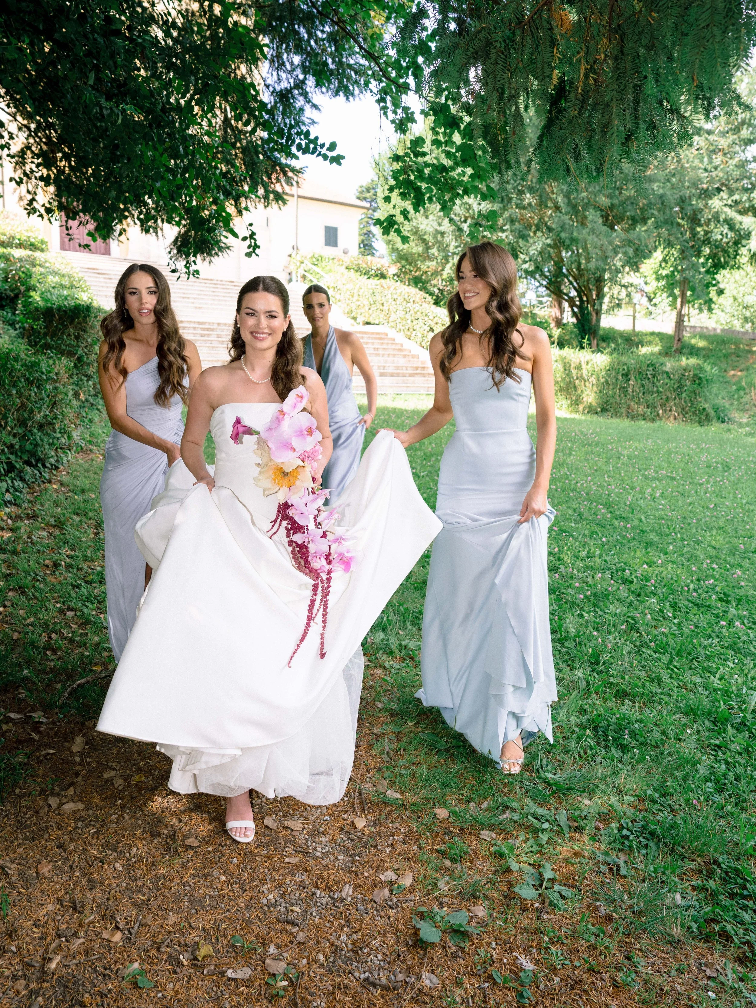 Four women in elegant dresses walking outdoors on a sunny day, surrounded by green trees and grass, with a building and stairs in the background.