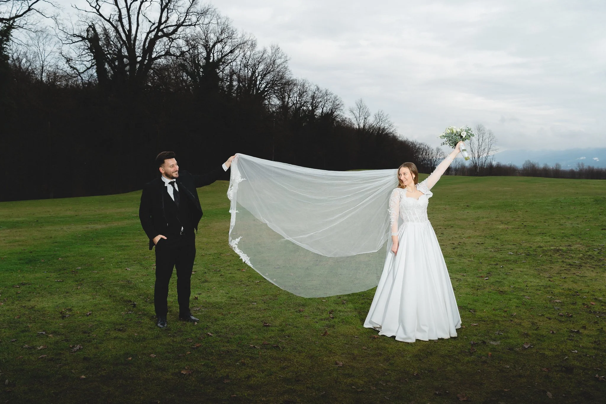 A bride and groom standing outdoors on a grassy field. The bride is wearing a white wedding dress with lace details, holding a bouquet, and raising her arm with joy. The groom is dressed in a black tuxedo, smiling at the bride, holding her veil.