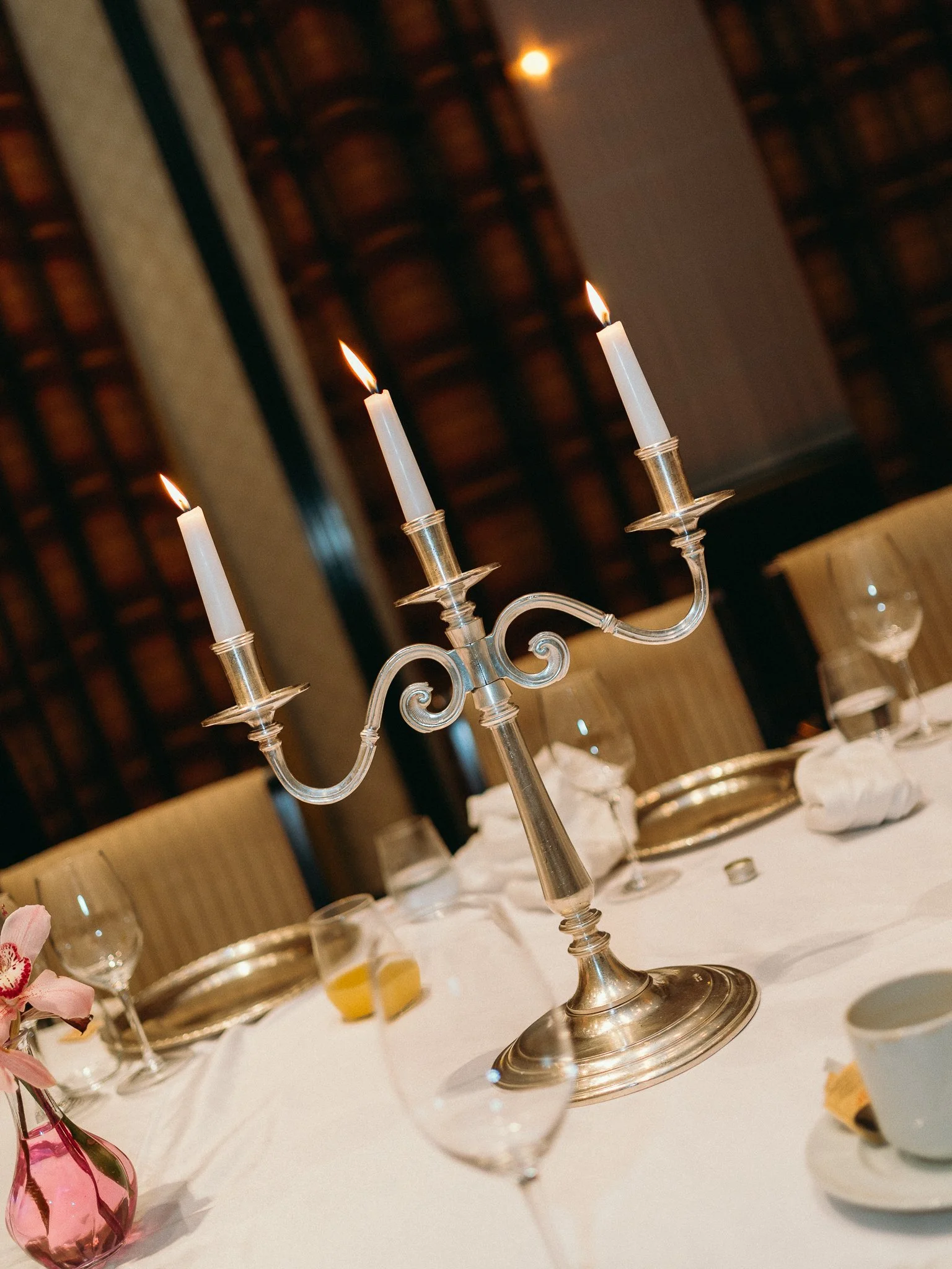 A silver candelabrum with three lit candles on a formal dining table with glassware and a small pink flower arrangement.