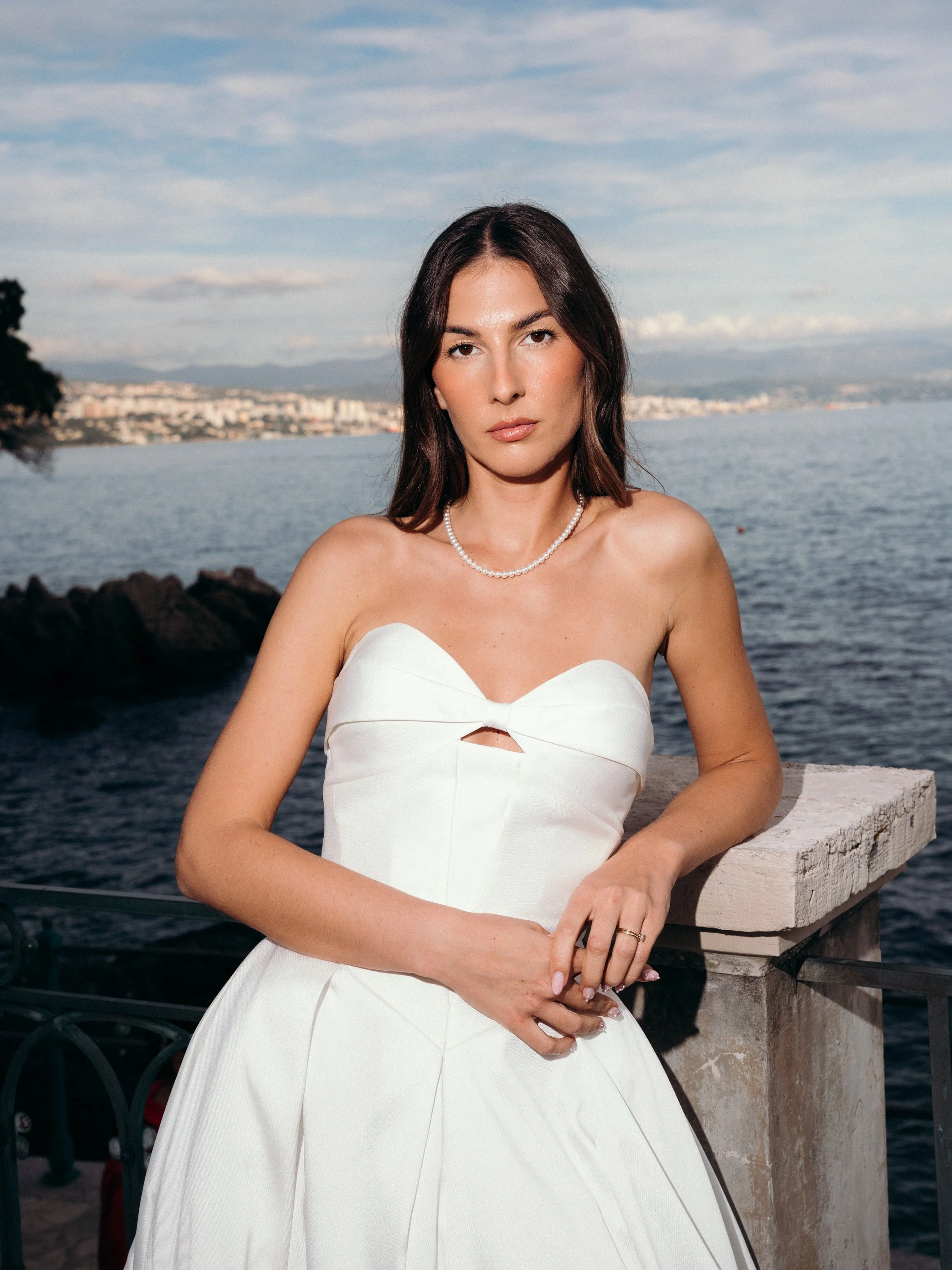 A young woman in a white strapless dress with a small cutout, standing by a stone railing near the water with a city shoreline in the background and a cloudy sky.
