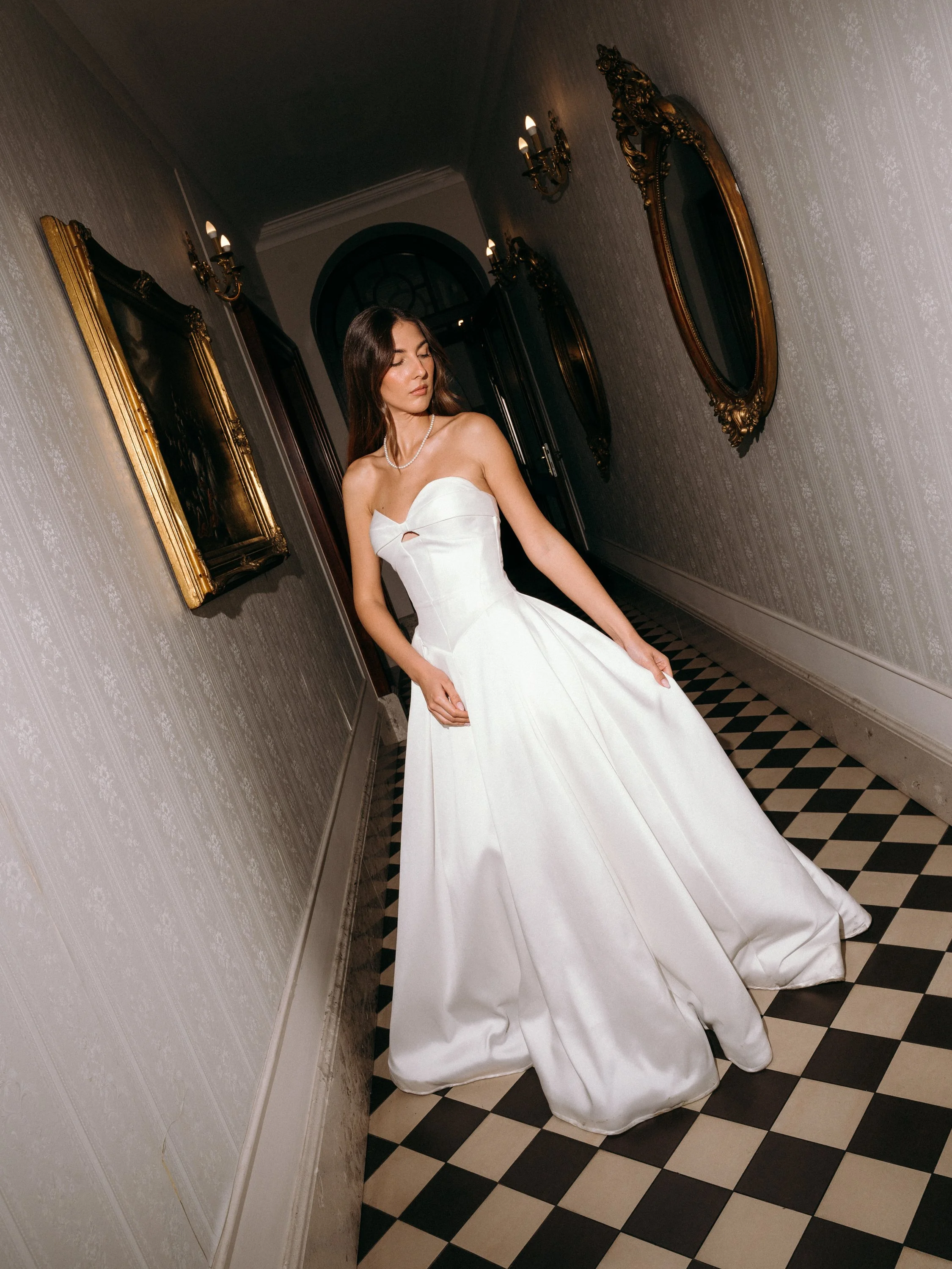 A woman in a strapless white wedding dress standing in a hallway with ornate gold-framed mirrors and wall sconces, black and white checkered floor, and neutrally patterned wallpaper.