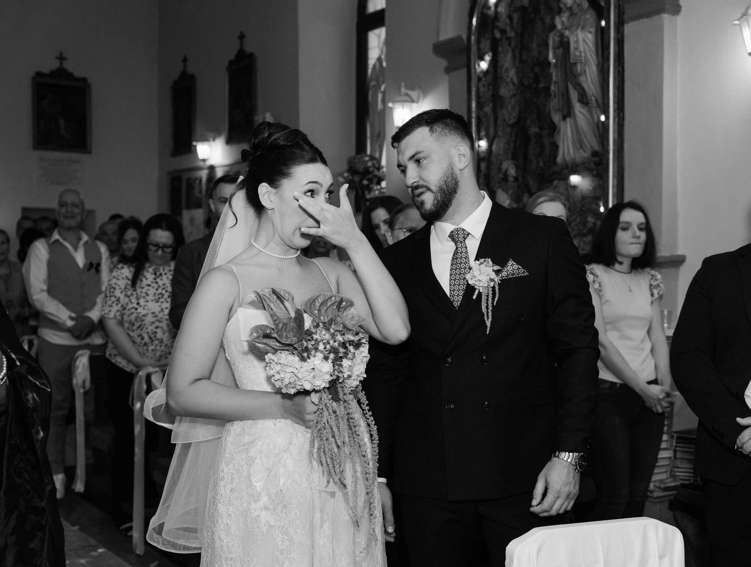 A bride and groom during their wedding ceremony, with the bride wiping her eye and the groom speaking to her, surrounded by guests in a decorated church.