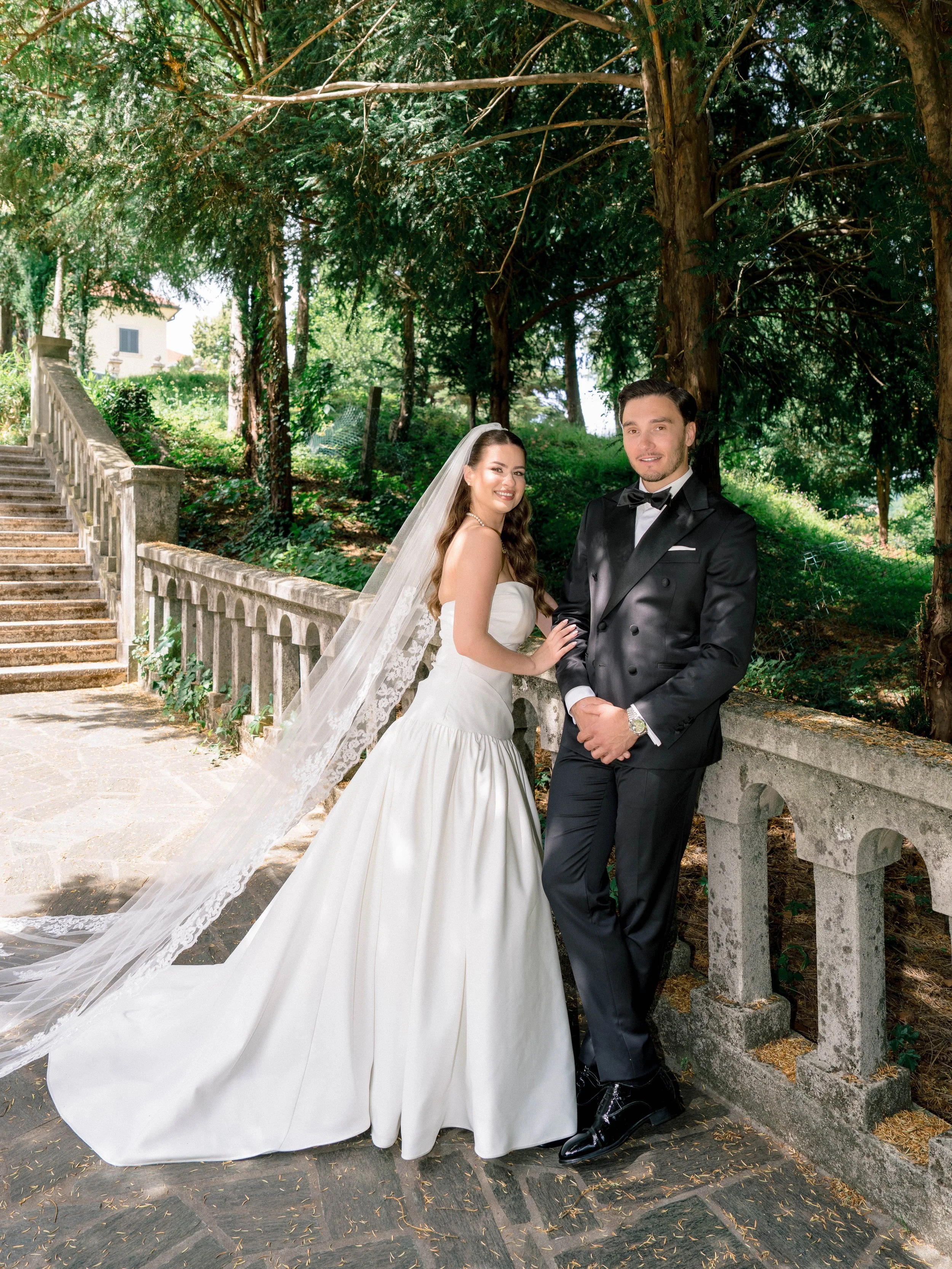 A newlywed couple posing on a stone bridge in a lush green park. The bride wears a white wedding gown with a long veil, and the groom wears a black tuxedo with a bow tie. Trees and stairs are visible in the background.