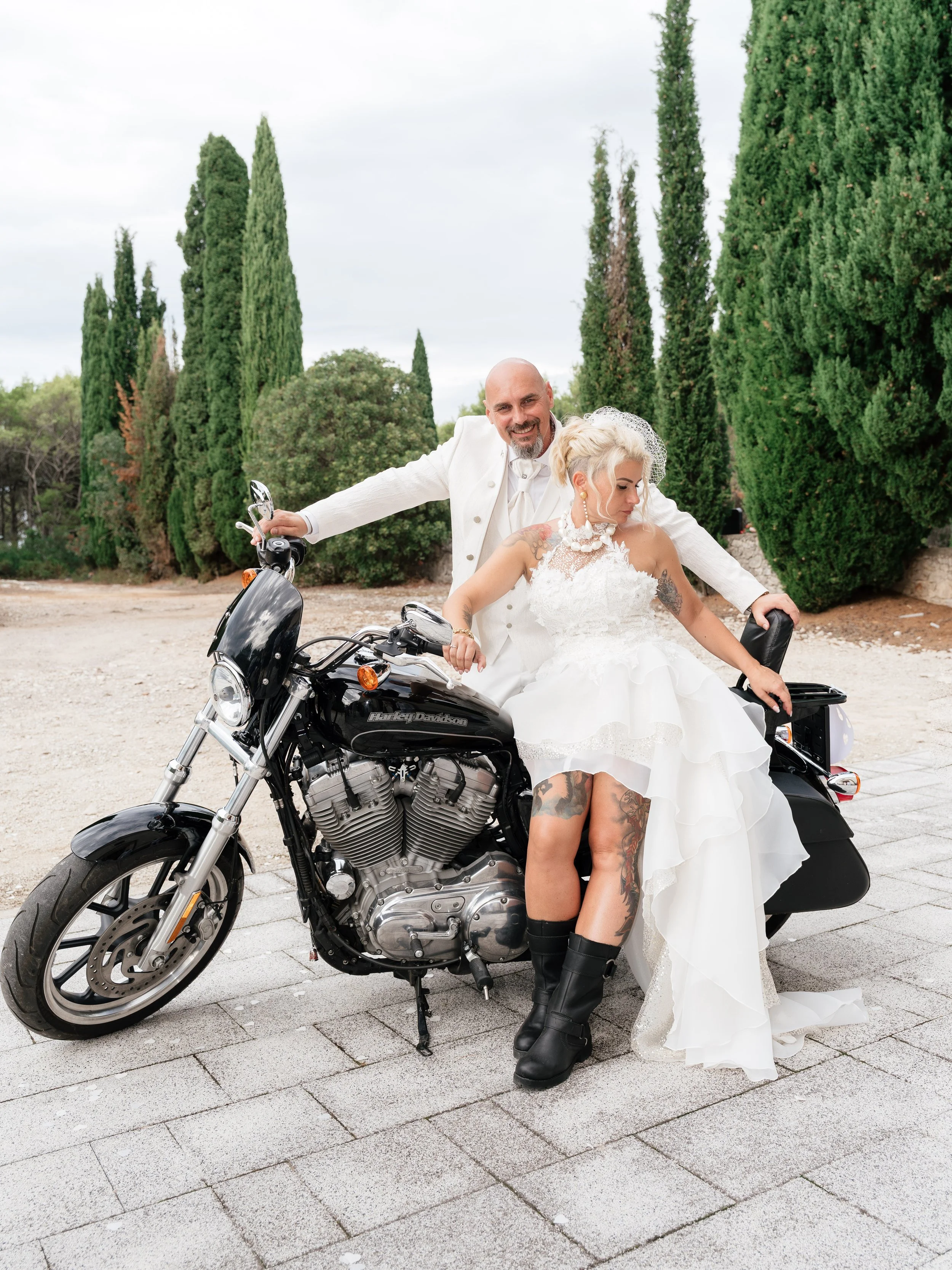 A man and woman in wedding attire sitting on a black Harley Davidson motorcycle outdoors, with tall green trees in the background.