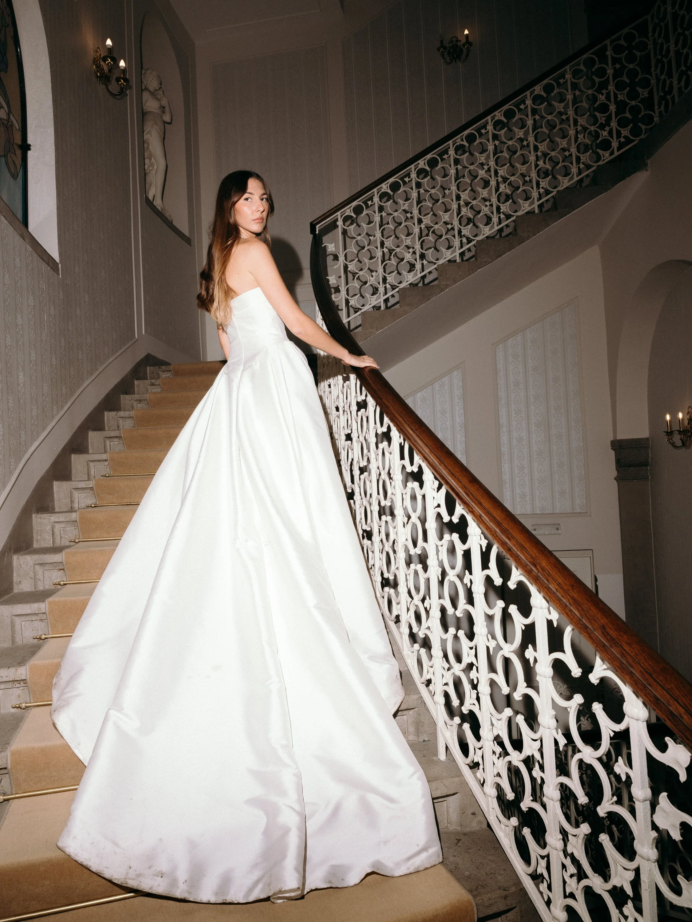 A woman in a white wedding dress standing on a staircase with ornate white railing and beige carpeted steps in a decorated indoor setting.