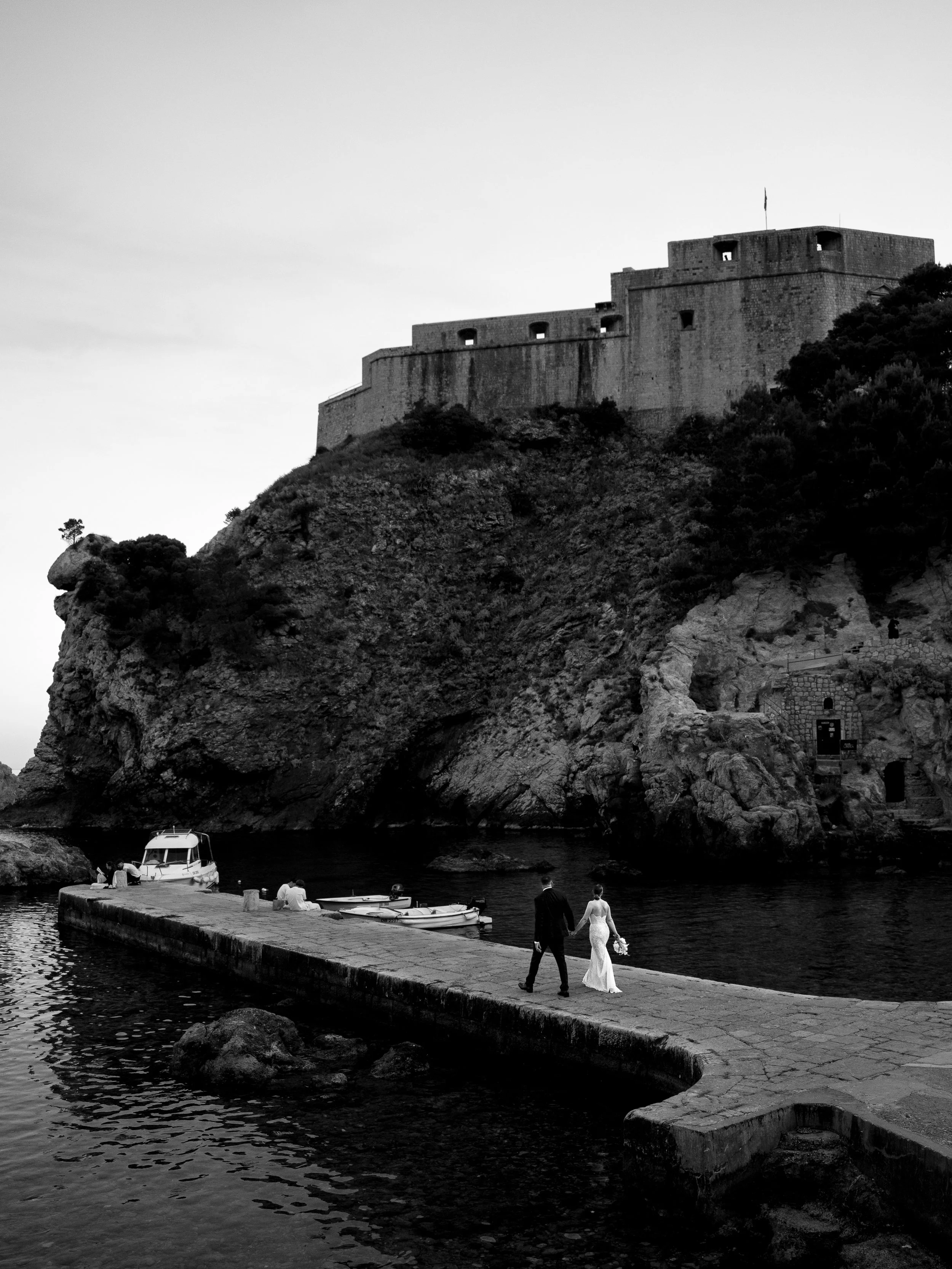 Black and white photo of a couple walking hand in hand along a rocky dock near a small boat and a larger boat, with a castle on a hill in the background.
