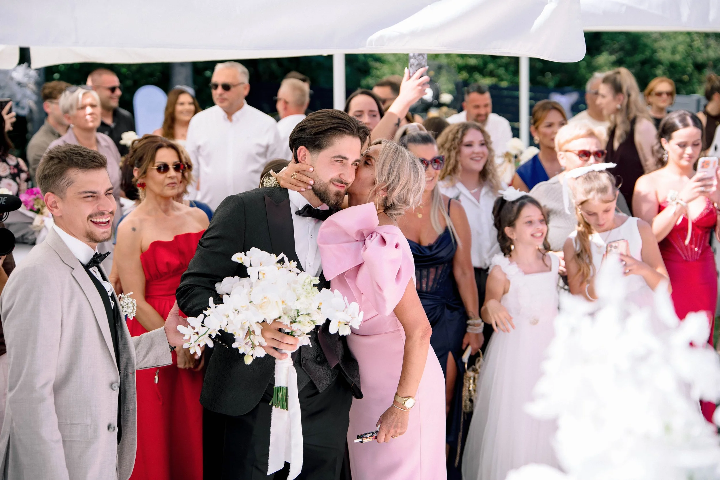A wedding celebration with a bride and groom, surrounded by guests, outdoors under large white umbrellas, with some guests taking photos and smiling.