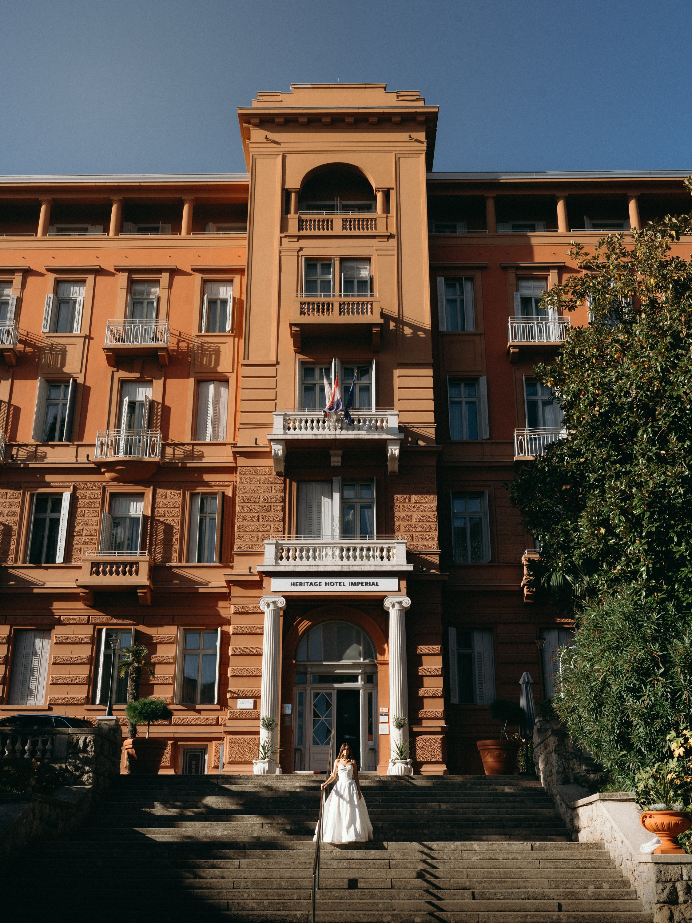 A woman in a white dress standing on the steps in front of the Heritage Hotel Imperial building, which has a peach-colored facade with multiple balconies and columns at the entrance, under a clear blue sky.