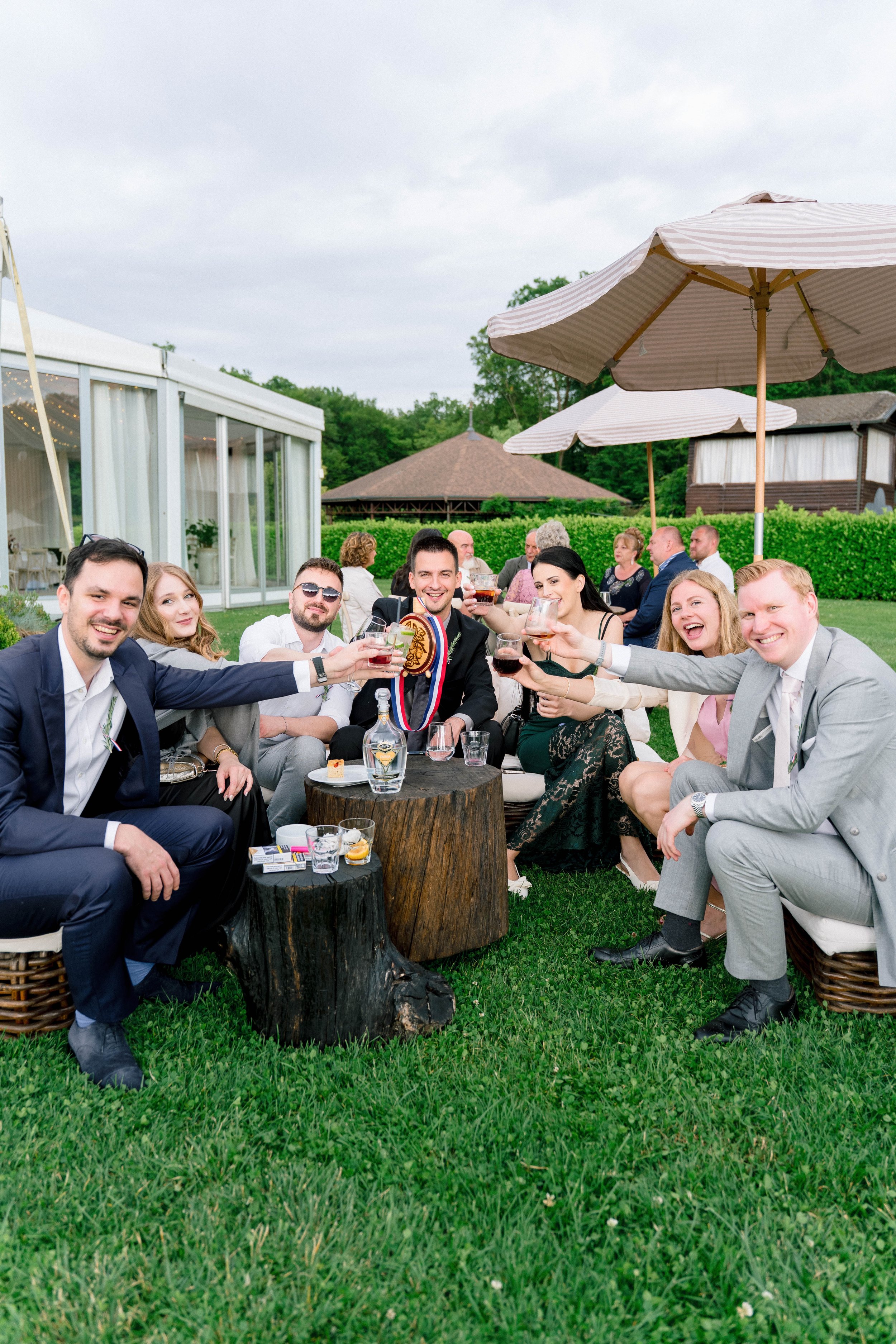 Group of people at an outdoor wedding celebration, raising glasses for a toast, sitting around a rustic wooden table with drinks and snacks, under large patio umbrellas, with a grassy area and a nearby glass-enclosed event space in the background.