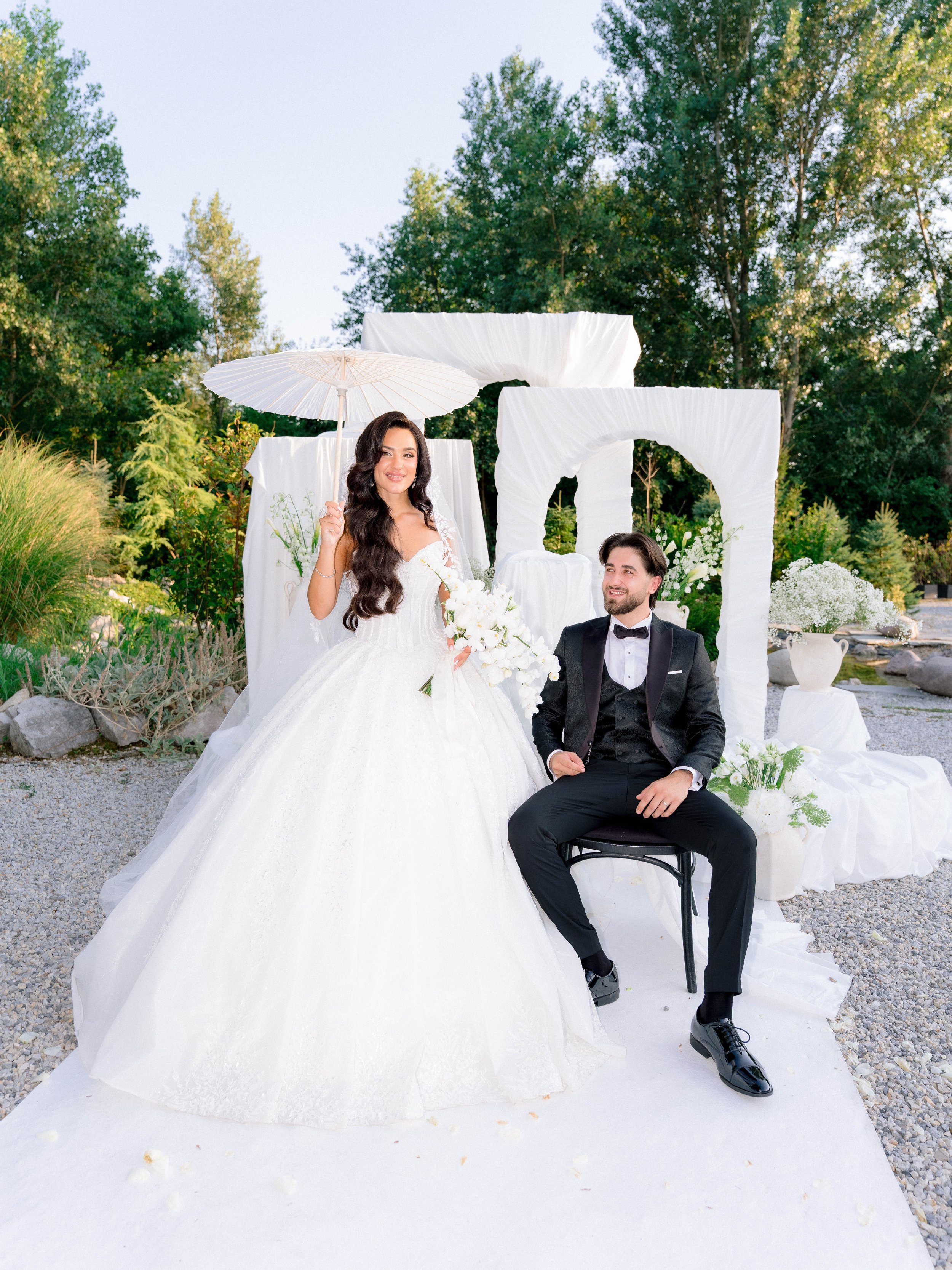 A bride and groom in wedding attire sitting outdoors with a white archway and greenery in the background. The bride is holding a bouquet and an umbrella, and the groom is smiling. The scene is decorated with white drapes and floral arrangements.