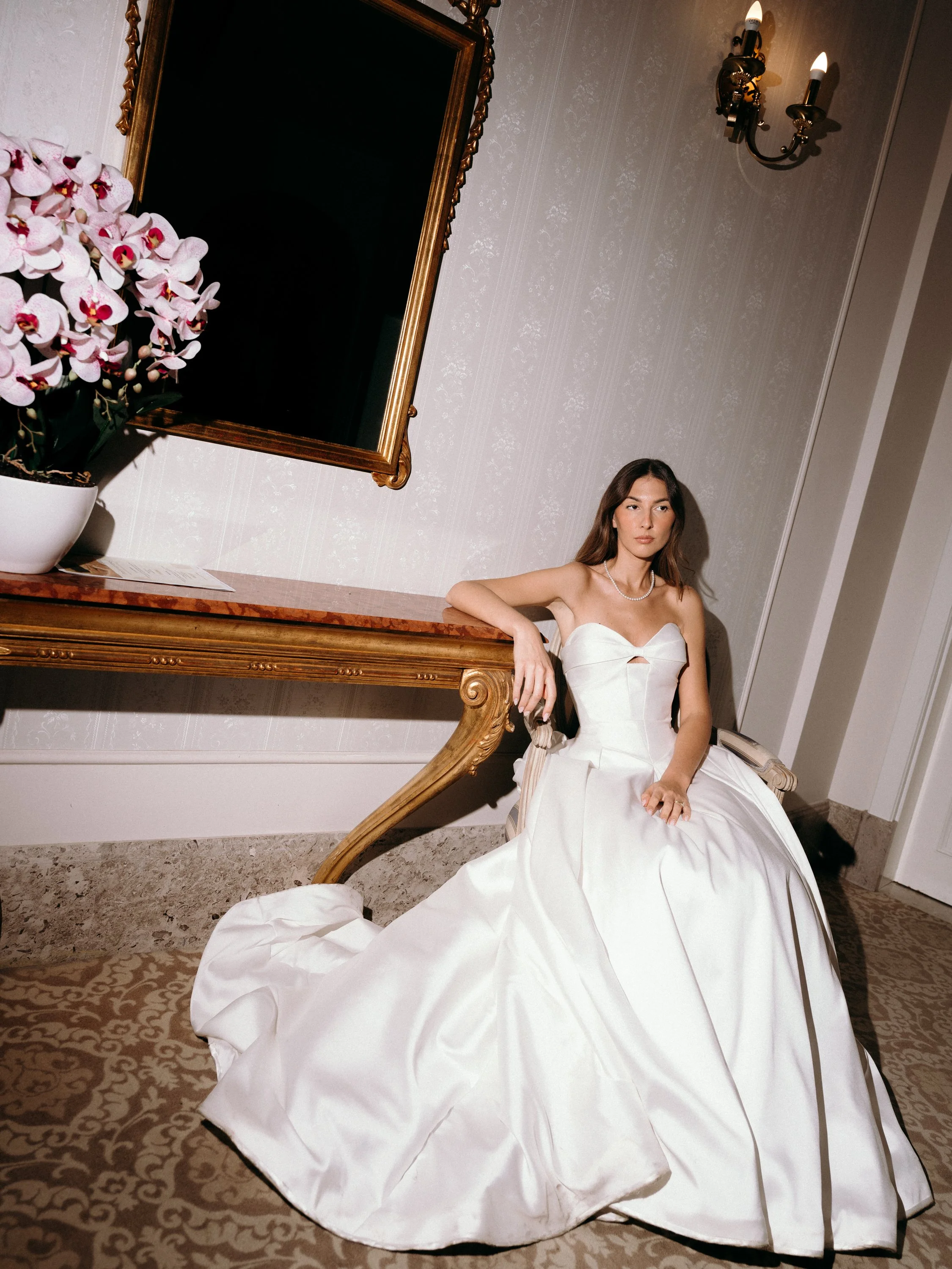 A woman in a white strapless wedding gown sitting on an ornate chair in a luxurious room with a marble-topped table, a large orchid plant, a mirror, and a wall sconce with lit candles.