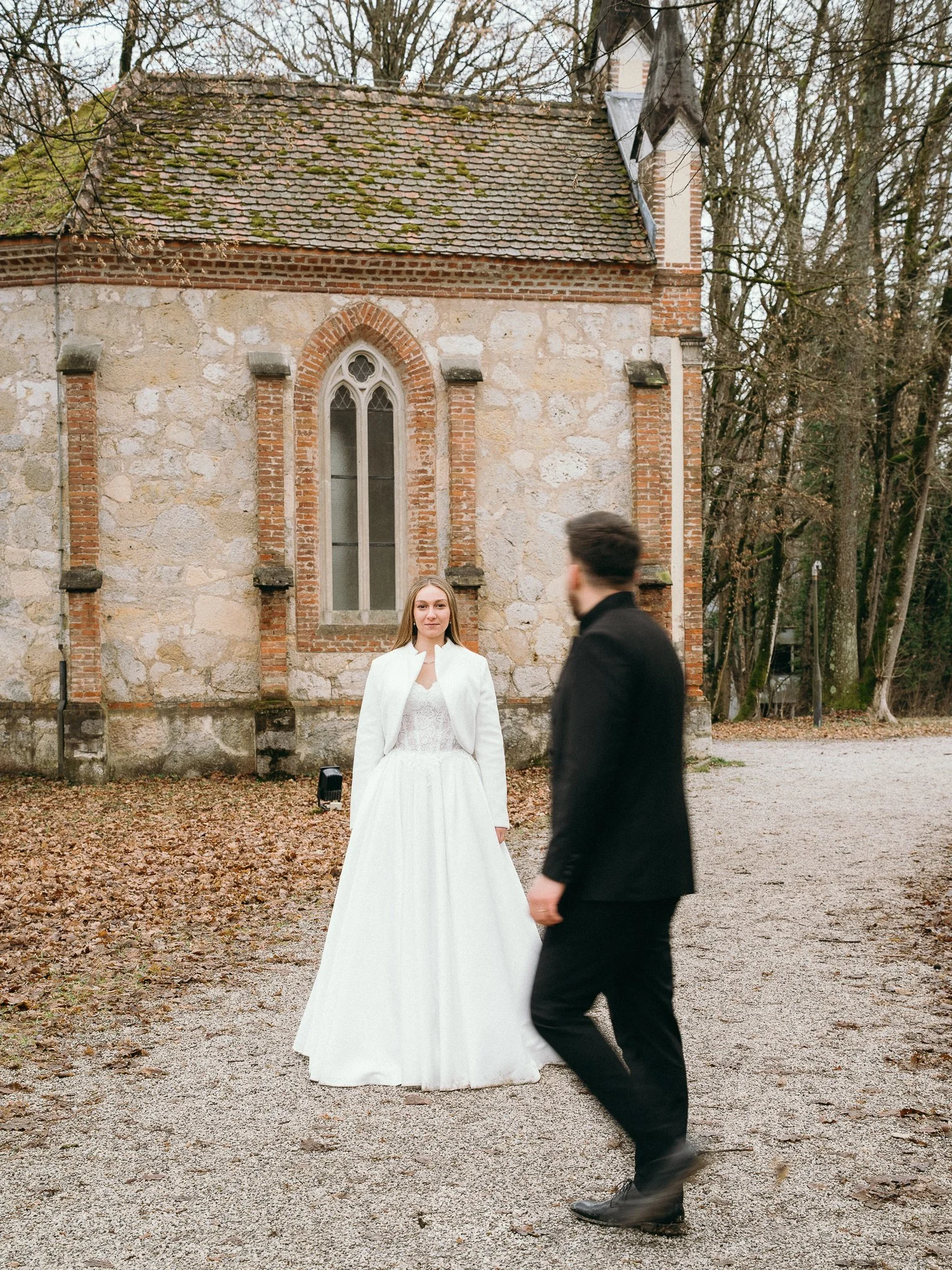 A bride in a white wedding dress and white jacket standing outdoors in front of a small stone church with a stained-glass window, while a man in black walks past.