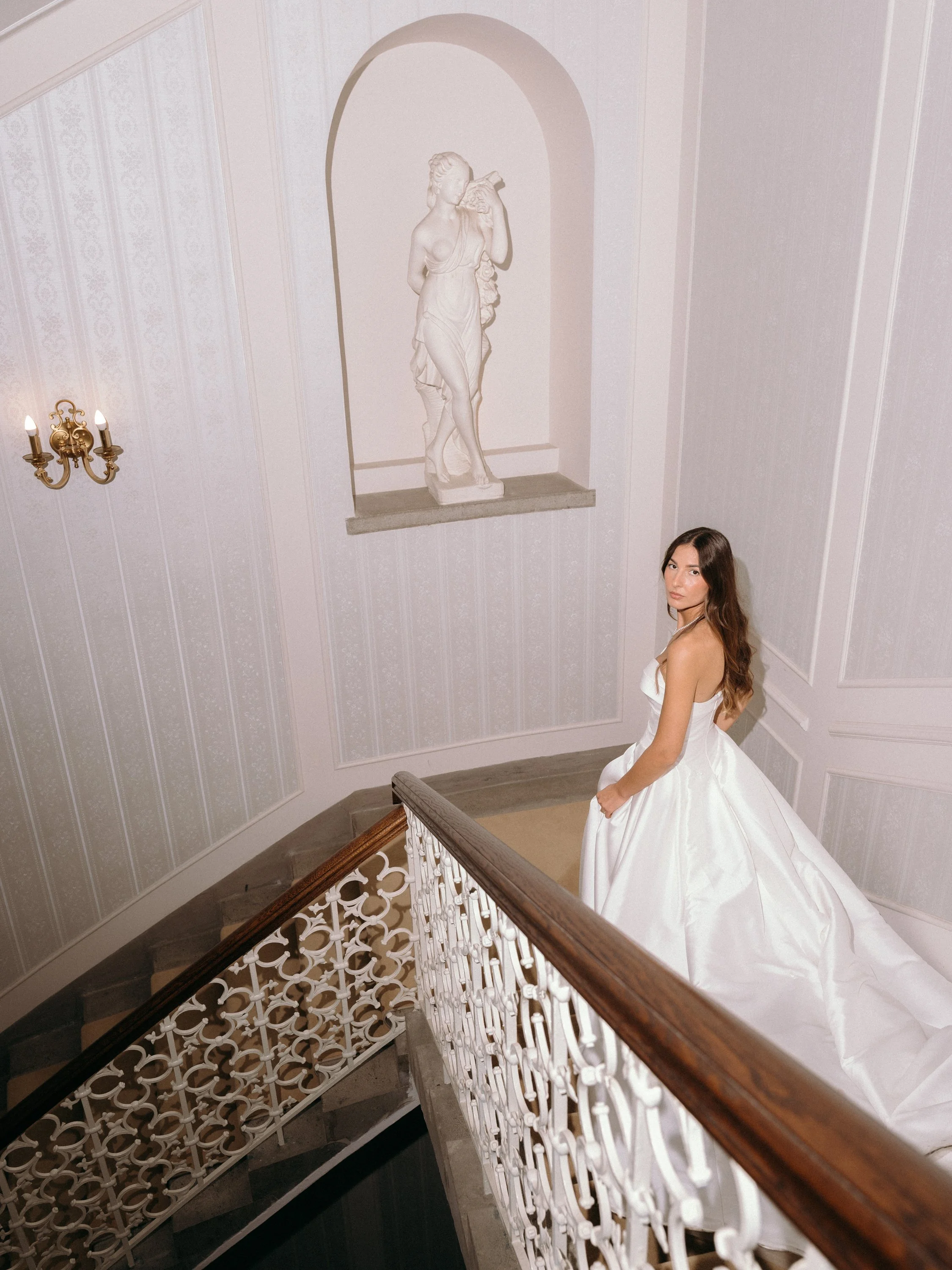 Woman in white gown standing on staircase near a wall with a classical statue in a niche