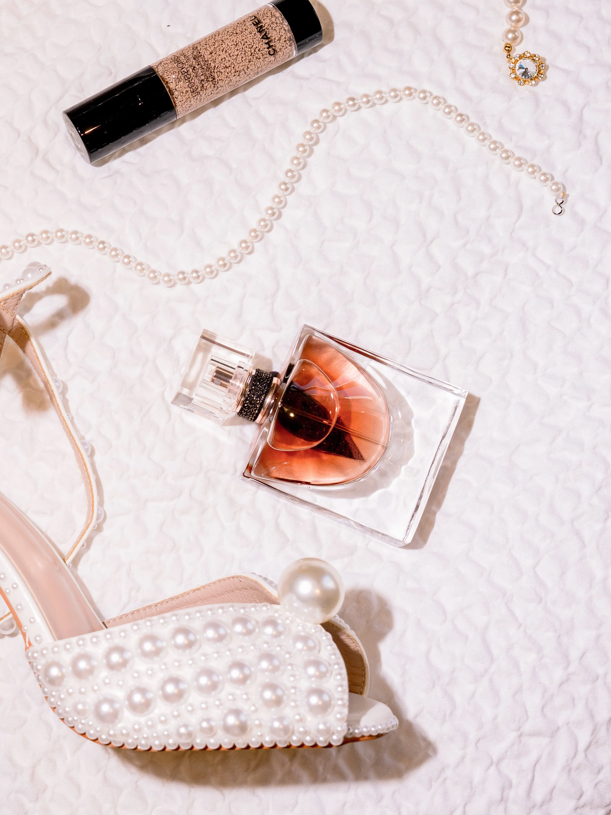 A pair of high-heeled shoes decorated with pearls, a pearl necklace, a perfume bottle, and a makeup foundation on a white textured surface.