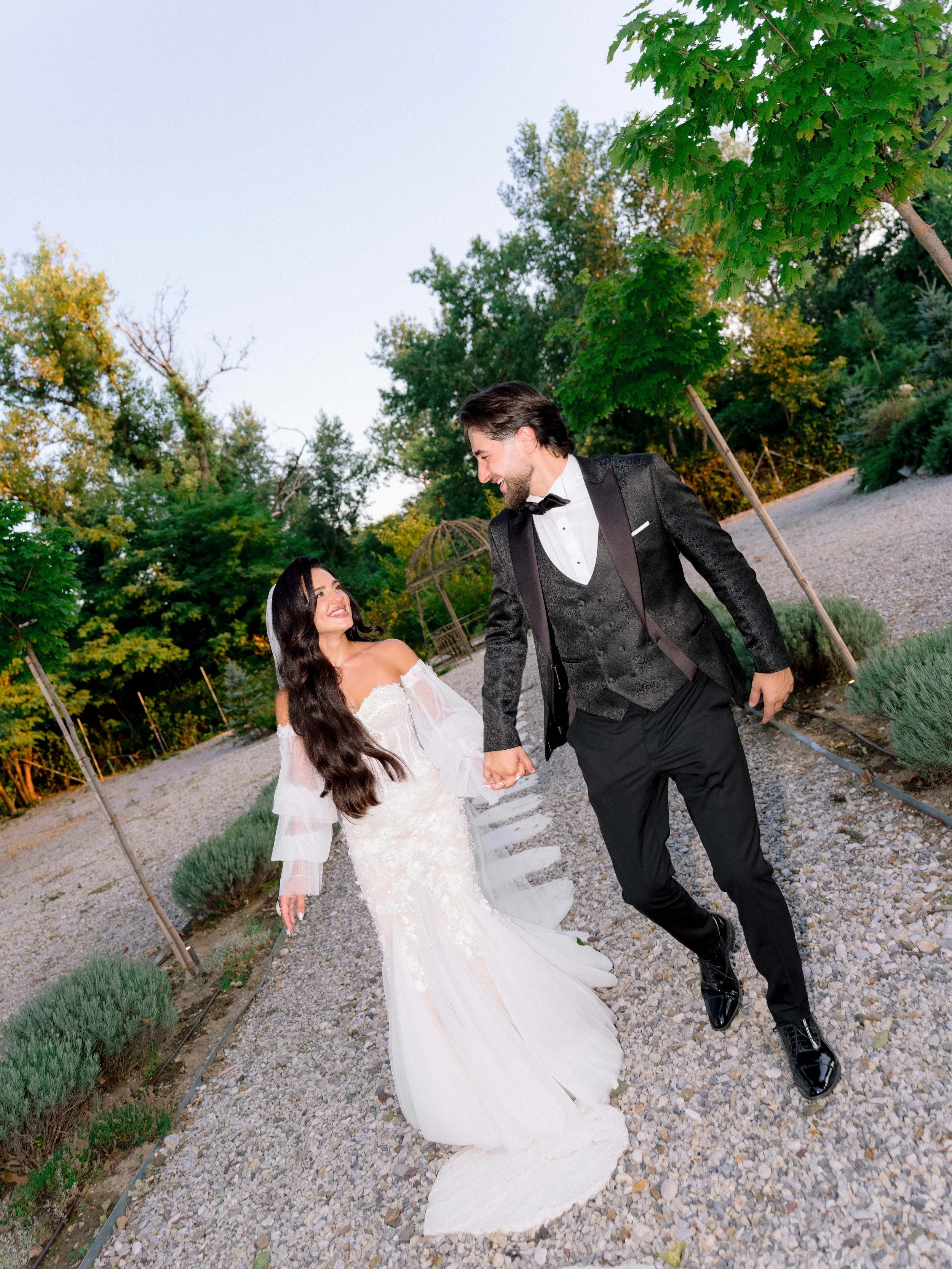 A newlywed couple holding hands and smiling while walking outdoors on a gravel pathway surrounded by green trees and plants. The bride is wearing a white wedding gown with lace details and the groom is dressed in a black tuxedo with a bow tie.