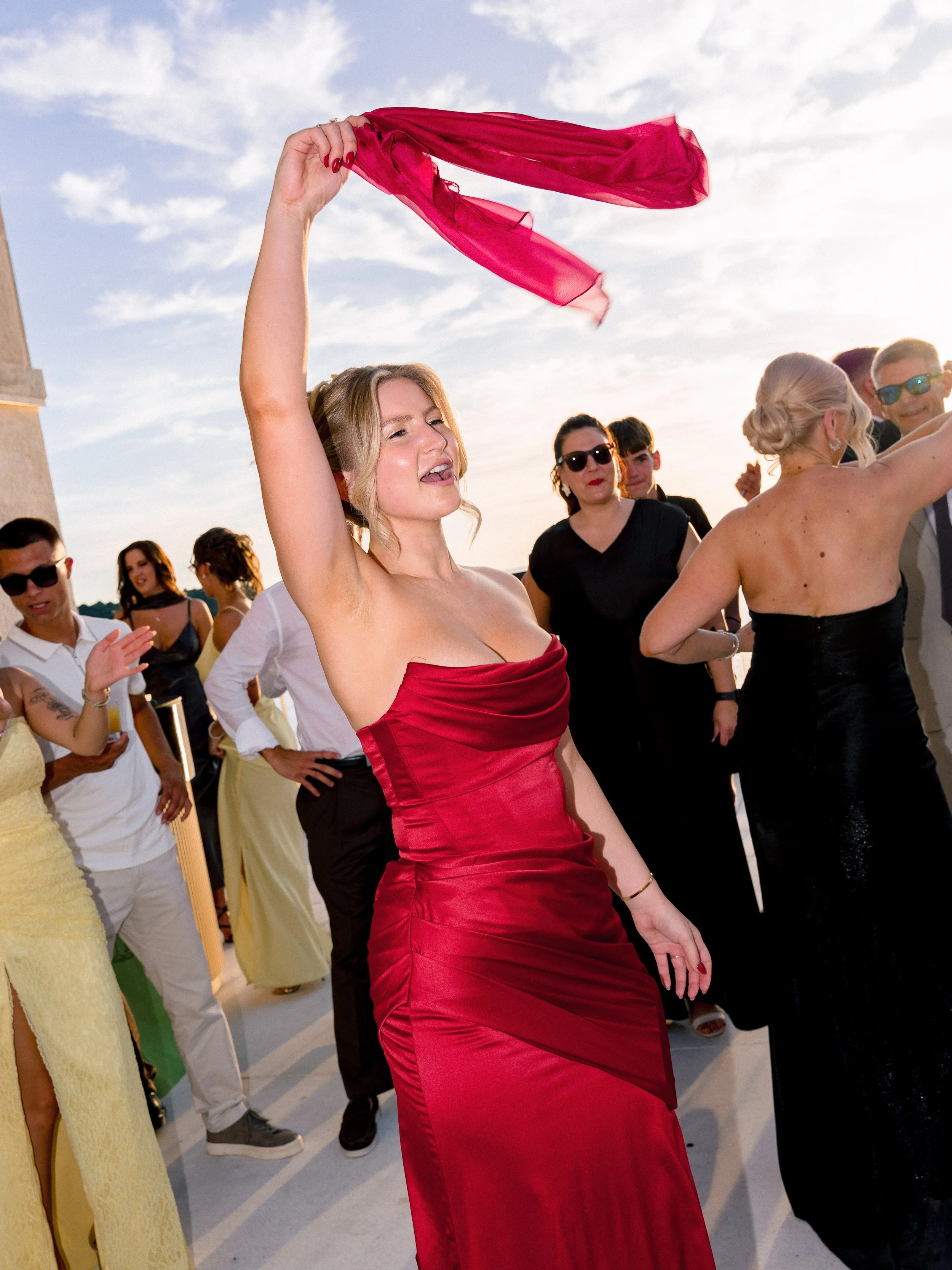 Woman in a red satin dress waving a red scarf at an outdoor party with many people around during sunset.