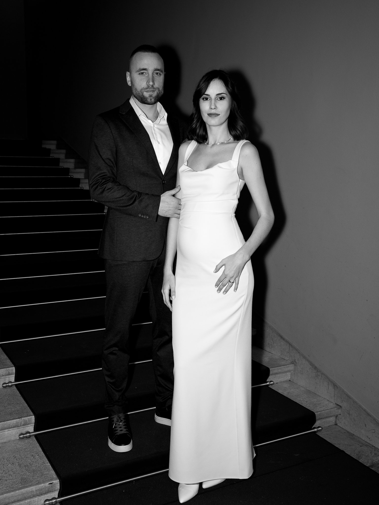 A man and woman standing together on a staircase, dressed in formal attire, with the man in a suit and the woman in a long gown, in a black-and-white photo.