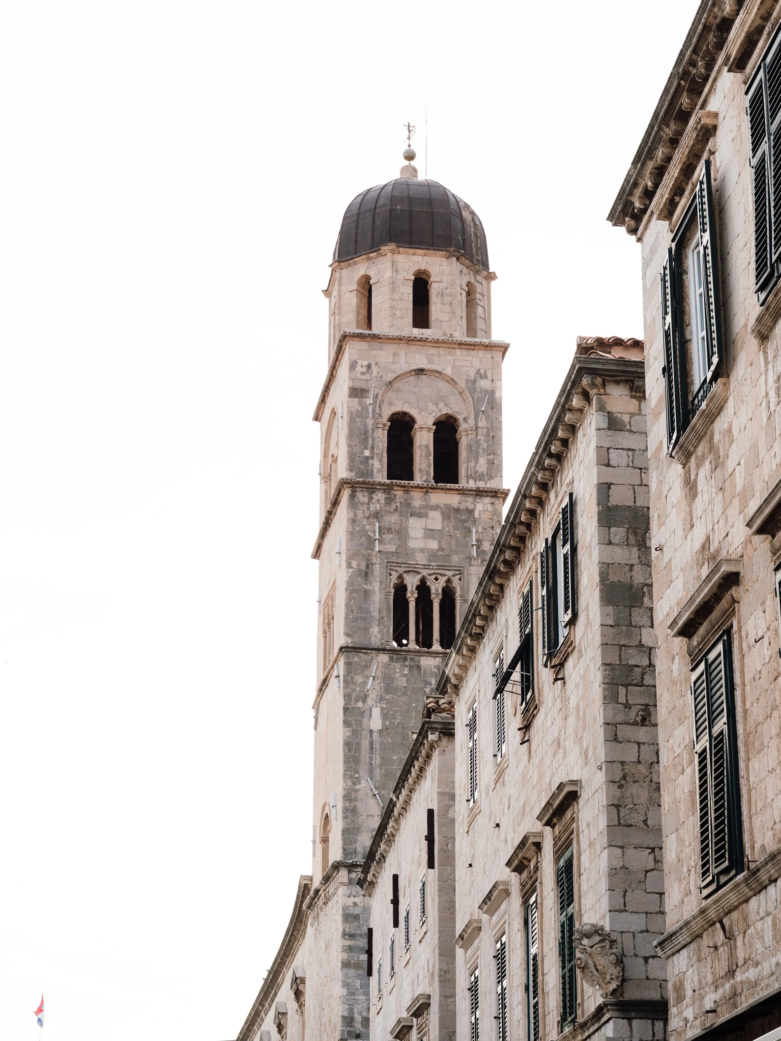 View of an old European stone building with a tall clock tower and black shuttered windows.