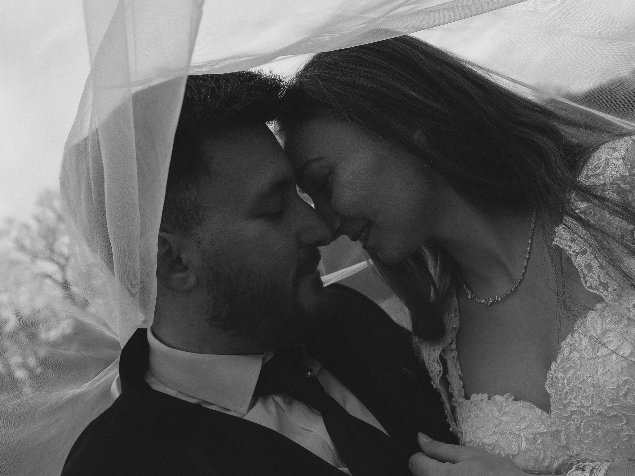 A black and white photo of a bride and groom close together, noses touching, under a wedding veil, with outdoor trees in the background.