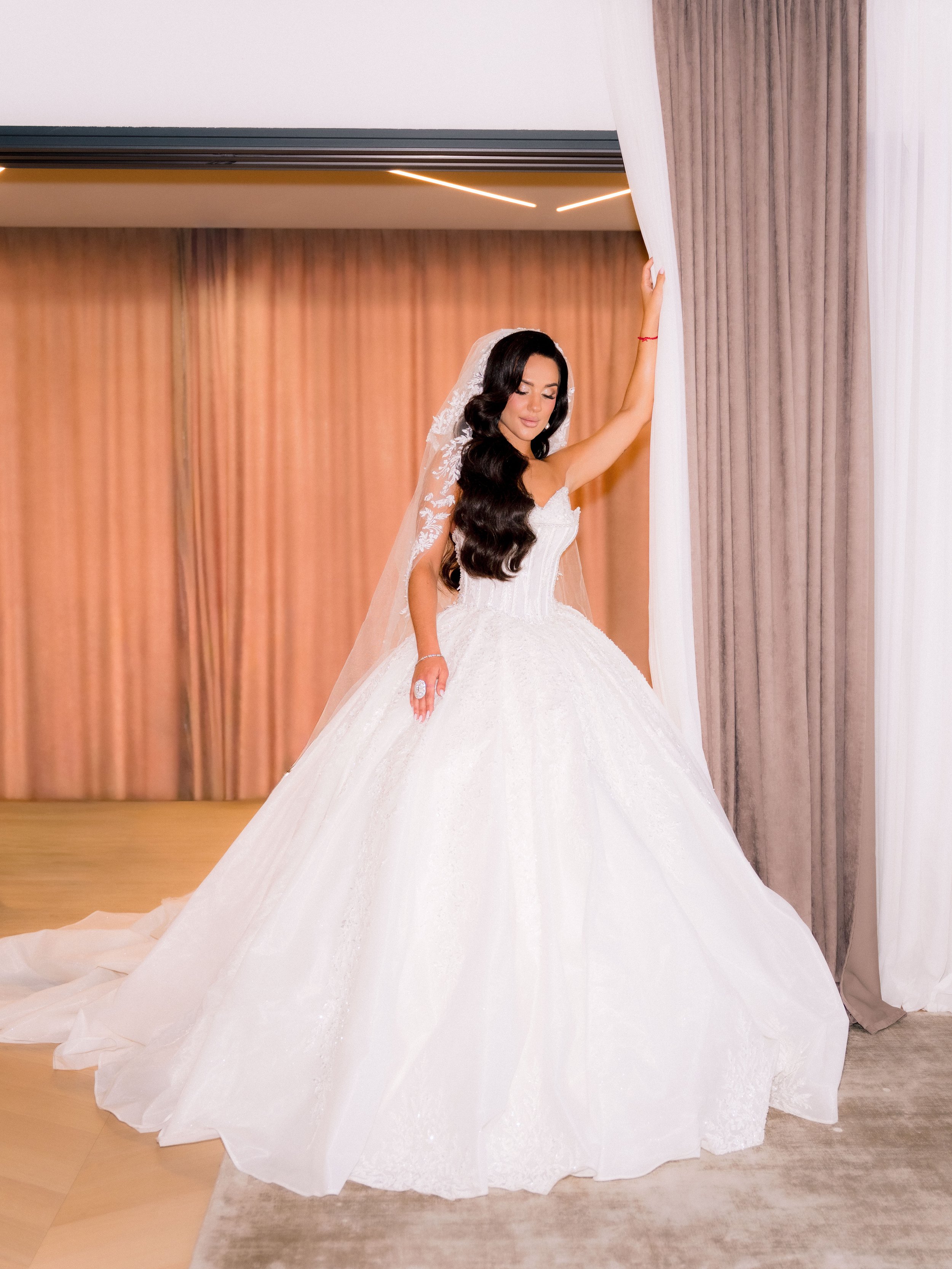 A woman in a white wedding gown holding back a curtain in a hotel or event hall.