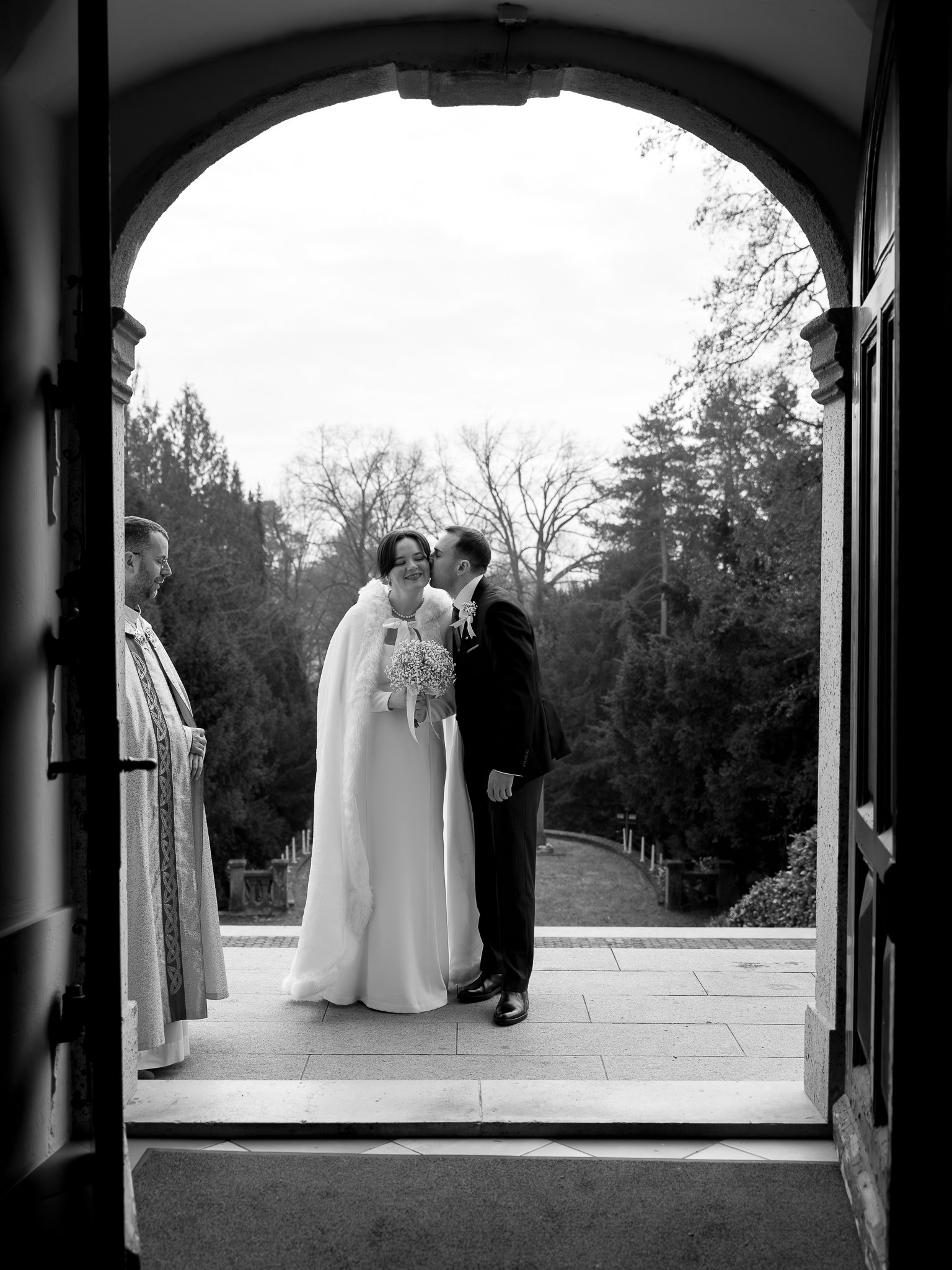 A black-and-white photo of a wedding scene taken from inside a building through an arched doorway. A bride and groom are standing outside on the steps, sharing a kiss. The bride is holding a bouquet, and both are dressed in formal wedding attire. A p
