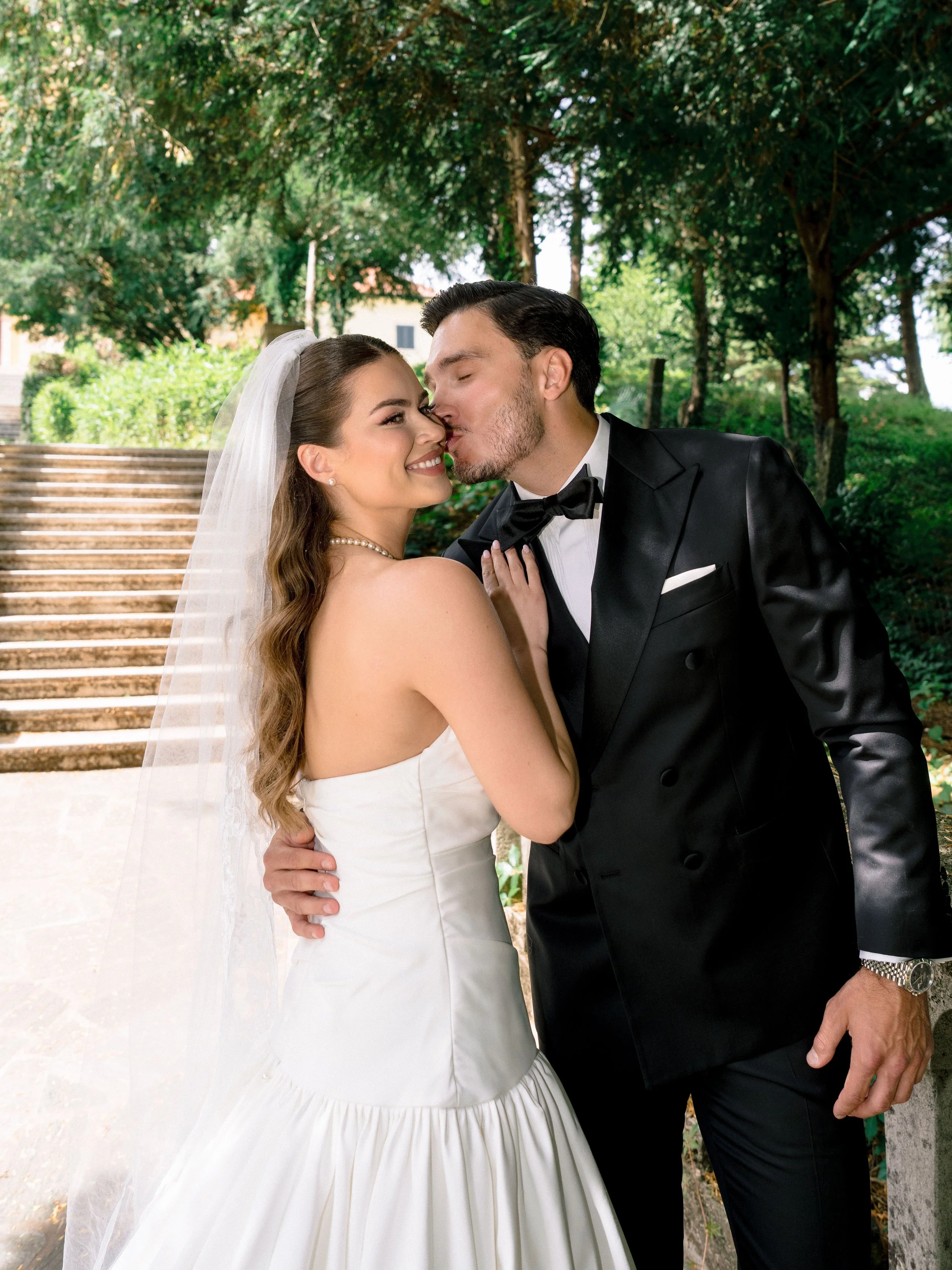 A bride and groom sharing a kiss outdoors during their wedding. The bride is wearing a white wedding dress with a veil and pearl necklace, while the groom is in a black tuxedo with a bow tie. They are standing on stone steps surrounded by green trees