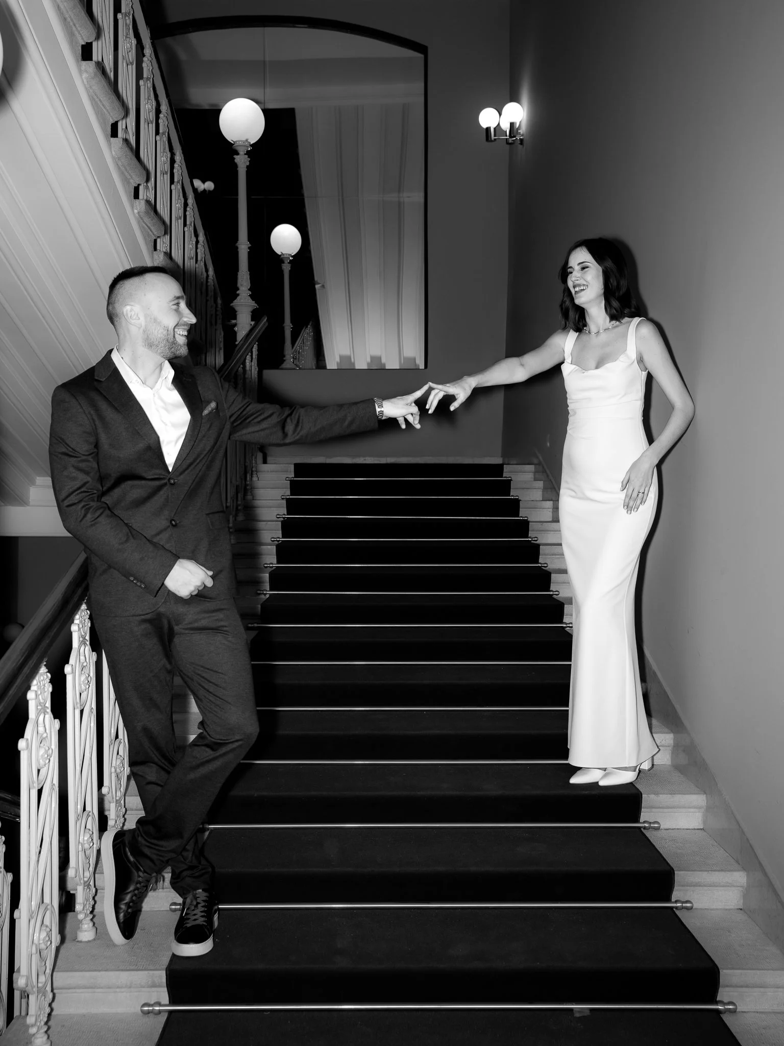 A man and woman on a staircase reaching out to each other, smiling. The man is in a dark suit and the woman is in a white dress. The setting appears to be elegant, with ornate railing and lighting.