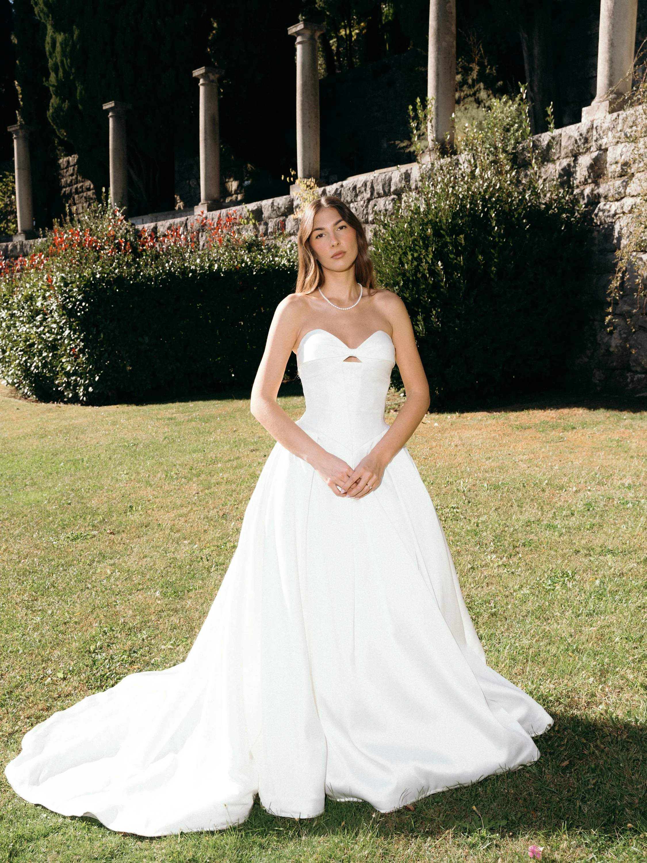 A woman in a white strapless wedding dress standing outdoors on a grassy lawn with bushes and stone columns in the background.