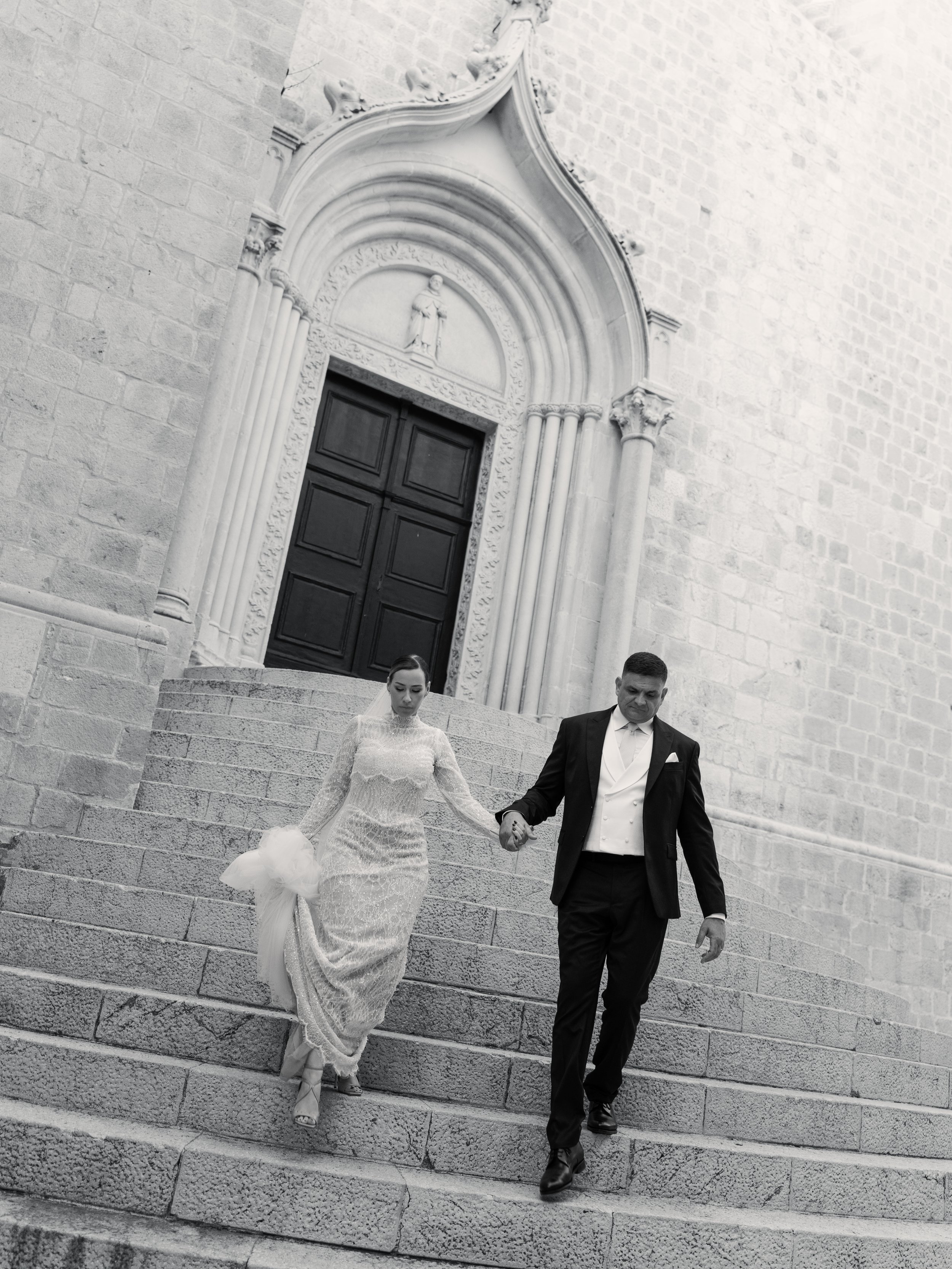 A bride and groom walking down stone steps outside a church. The bride is wearing a lace wedding dress and holding a bouquet, and the groom is in a tuxedo with a white shirt and tie. They are holding hands.