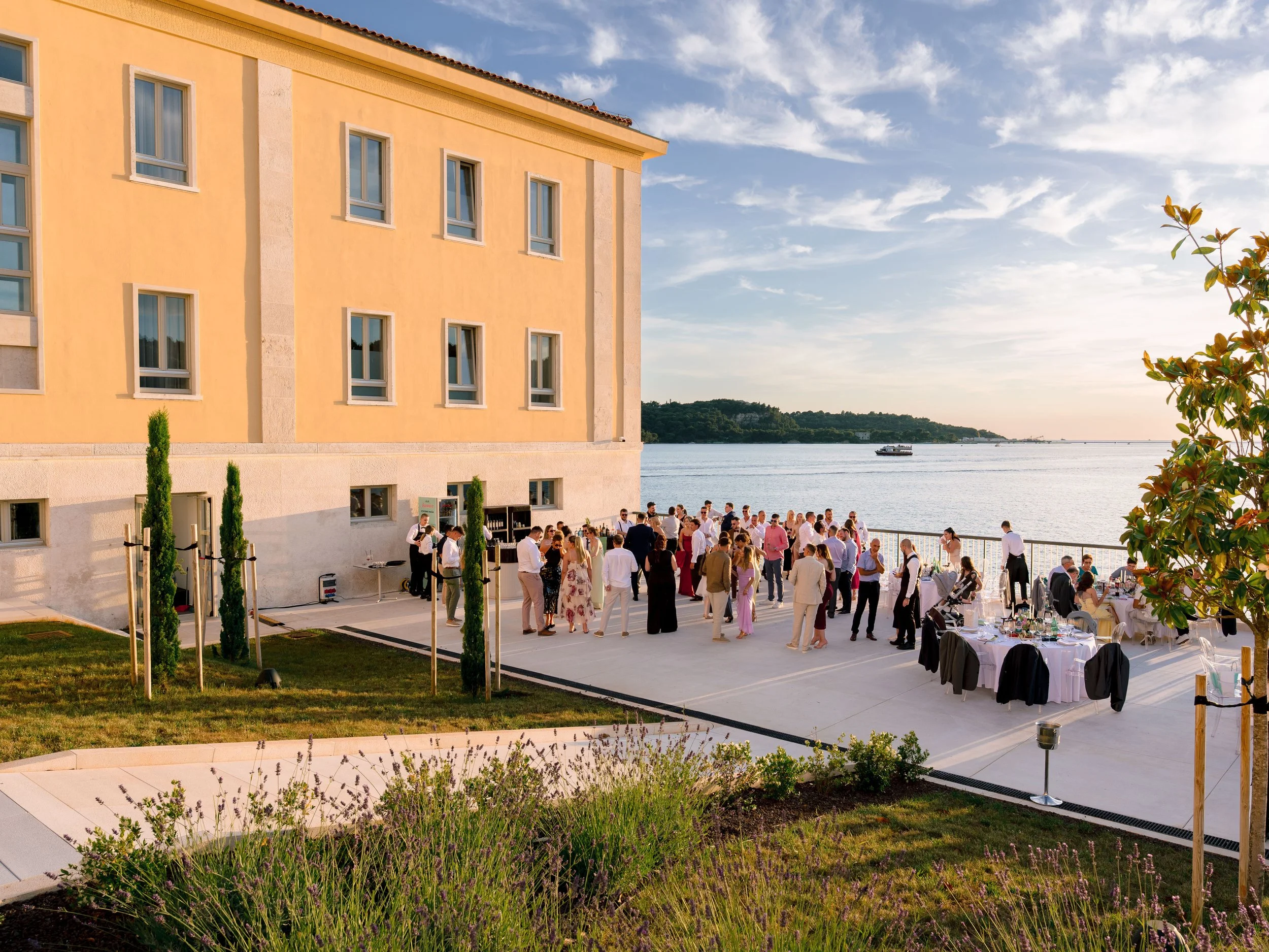 An outdoor wedding reception area by the water with guests gathered around tables, a yellow building on the left, and a boat on the water under a partly cloudy sky.