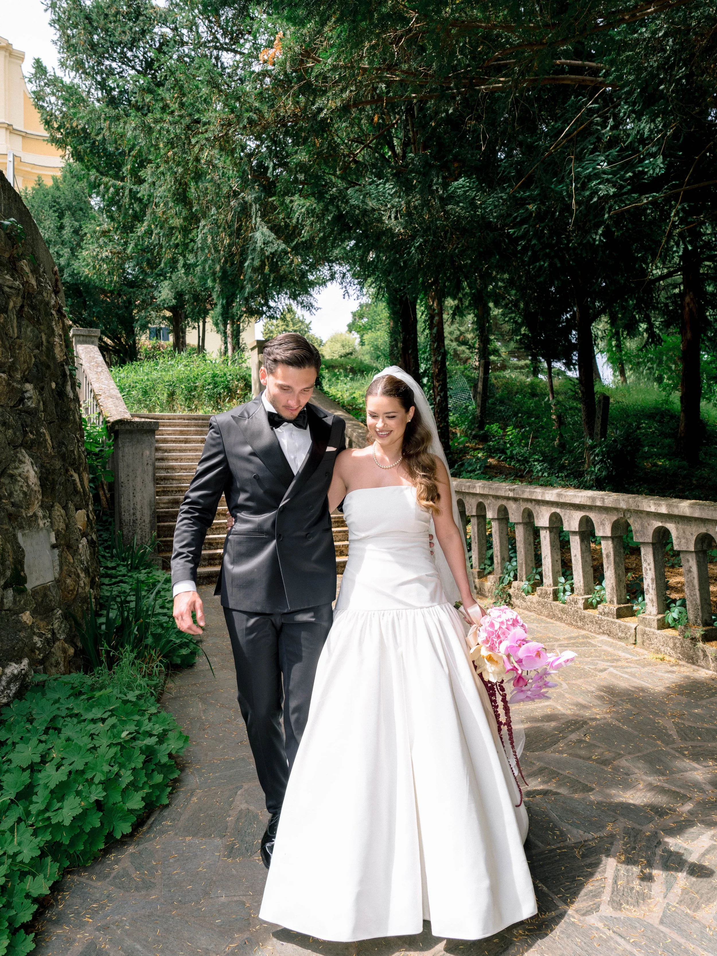 A newlywed couple walks outdoors on a stone path surrounded by lush green trees and plants. The groom wears a tuxedo and the bride is in a white wedding dress holding a bouquet of pink and white flowers.