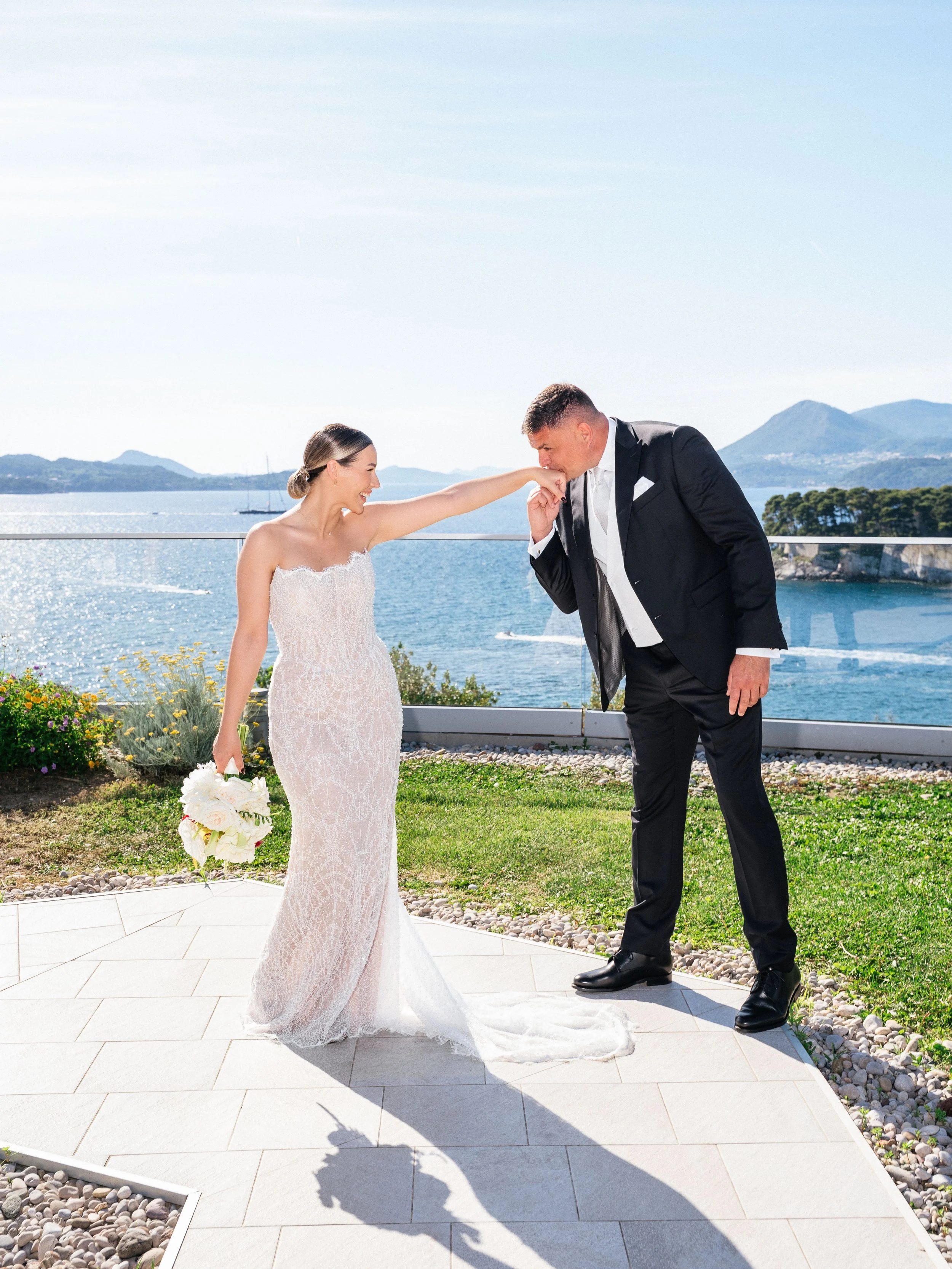A bride and groom share a romantic moment by the water with mountains in the background in Dubrovnik, Croatia. The bride in a lace wedding gown holds a bouquet and playfully touches the groom's nose, who is dressed in a black tuxedo.
