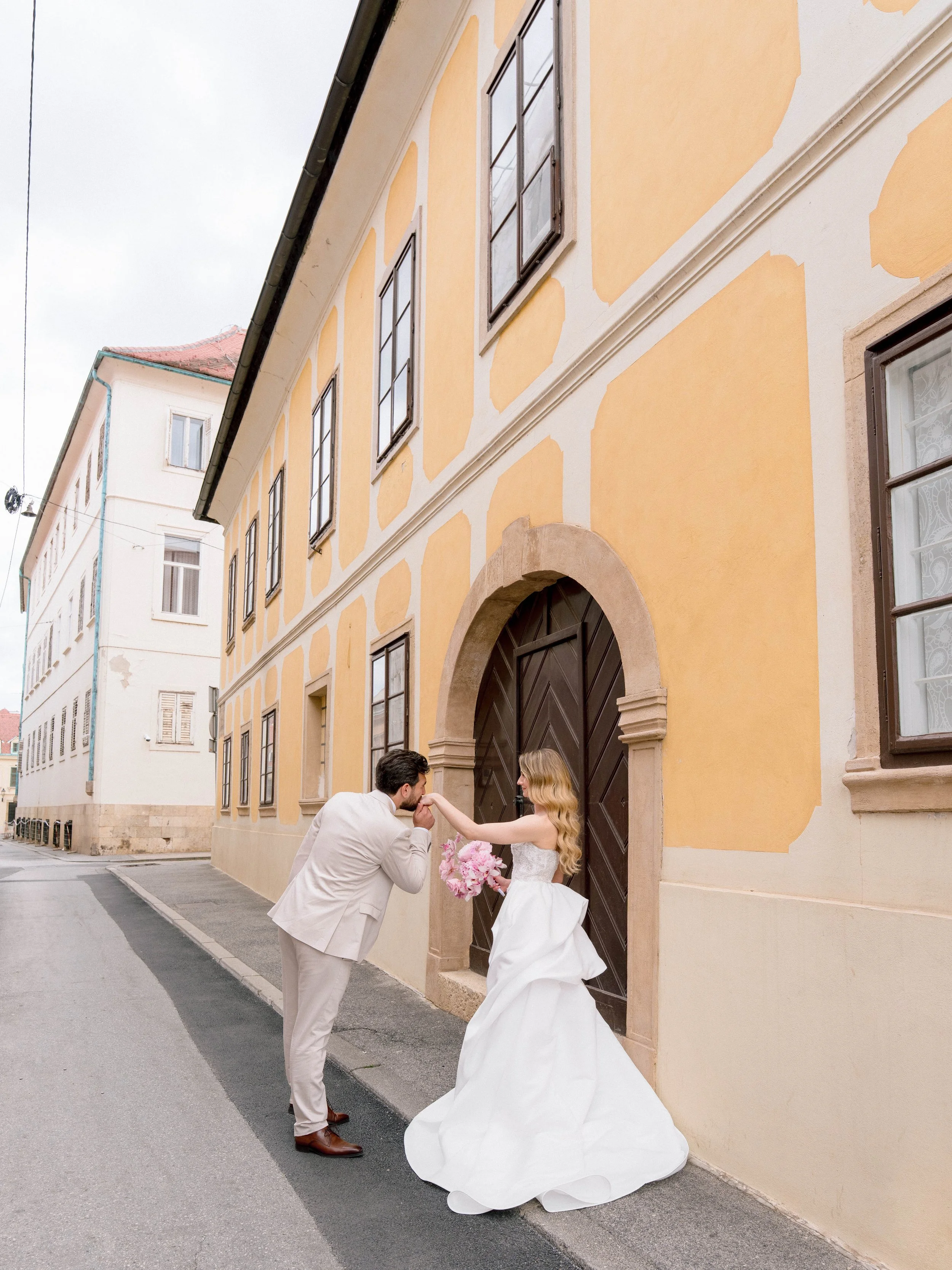 A newlywed couple is standing on a street outside a yellow building with black doors and windows. The groom is kneeling and kissing the bride's hand, who is holding a bouquet of pink flowers. The bride is wearing a white wedding gown, and the groom i