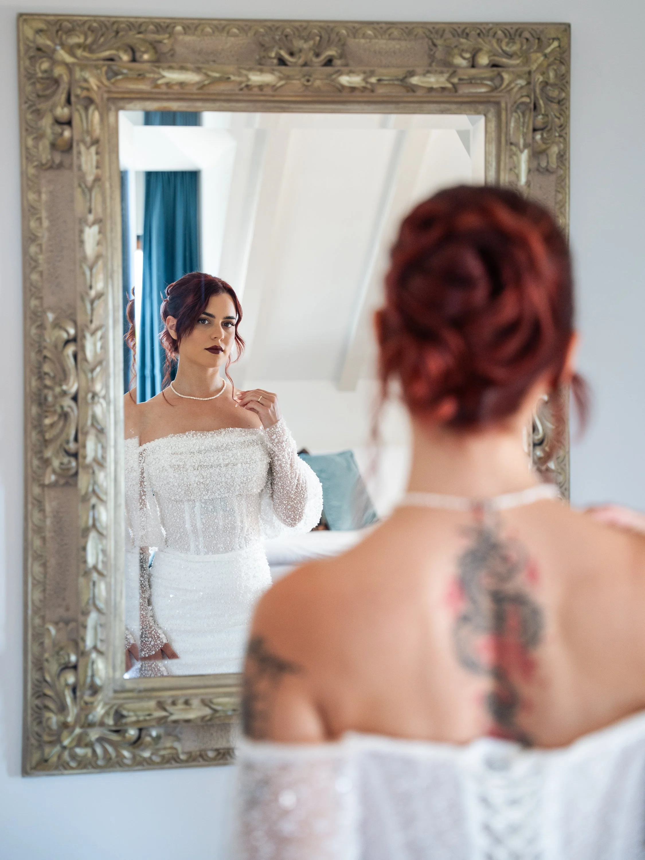 A woman with red hair styled in an updo looks at herself in a decorated mirror, wearing a white wedding dress with off-the-shoulder lace sleeves, necklace, and lipstick, with a tattoo visible on her back.