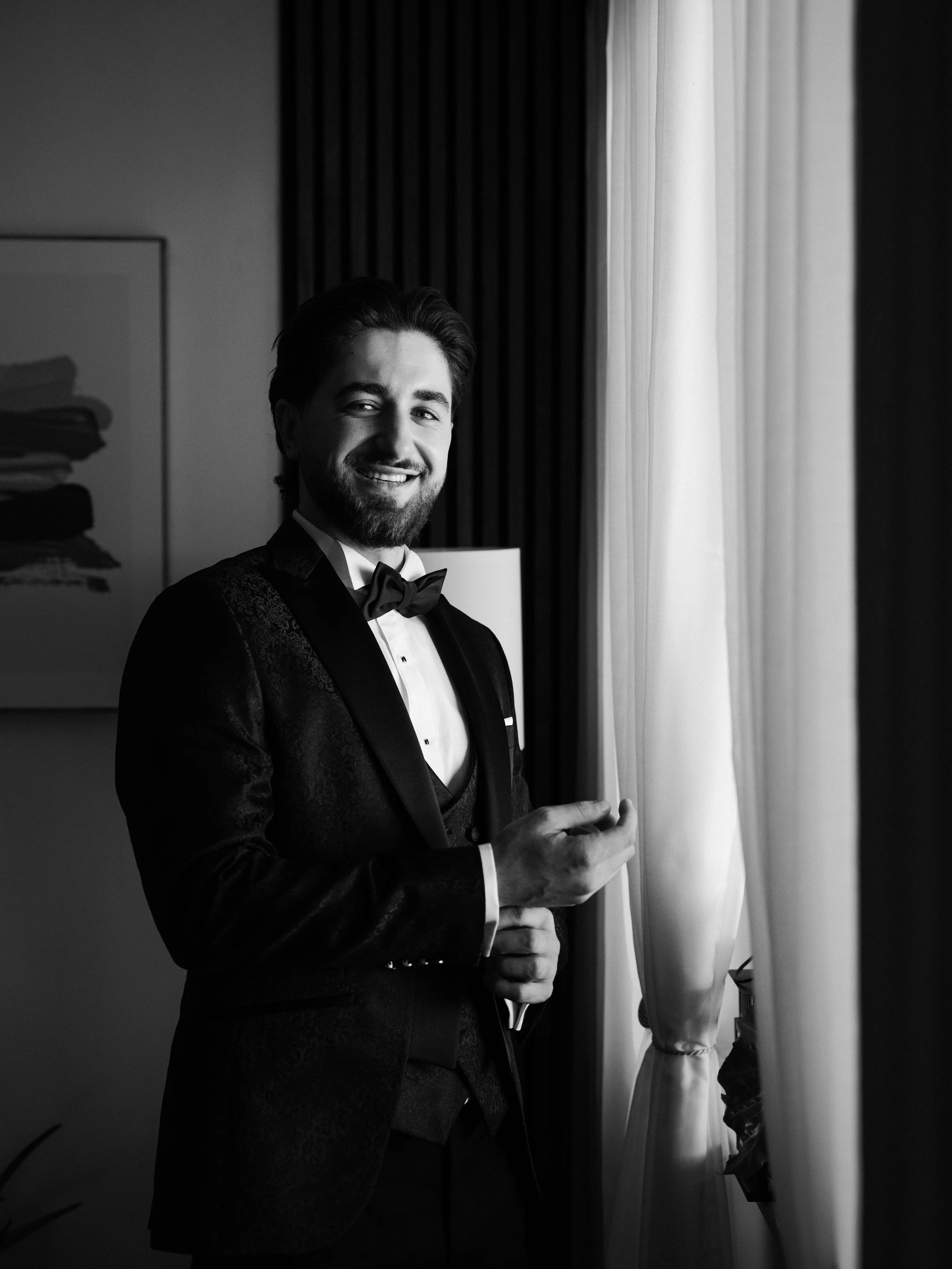 A man dressed in a tuxedo and bow tie is smiling and standing near a window with curtains, in a black-and-white photo.