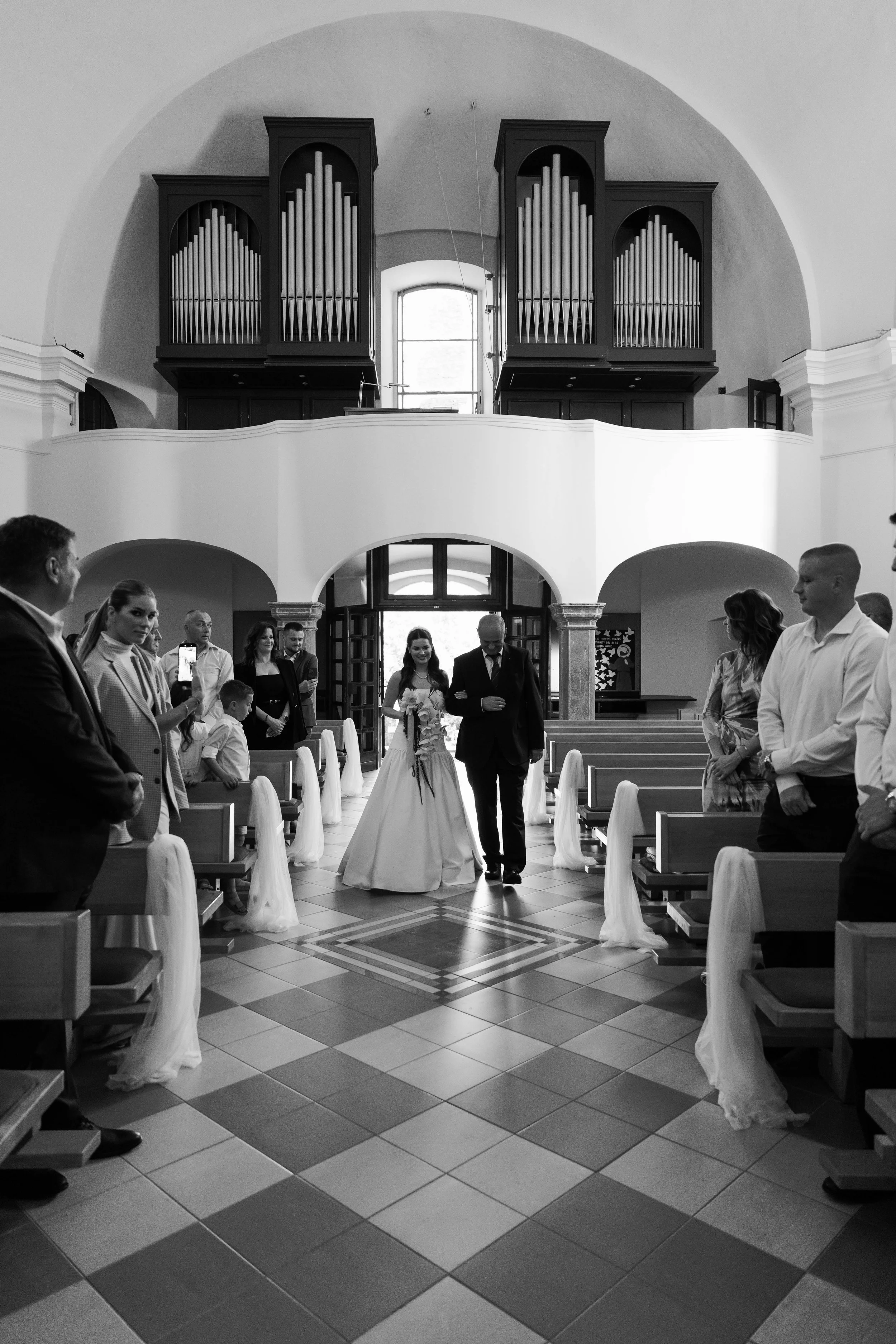Black and white photo of a wedding ceremony inside a church with pews decorated with white fabric. A bride in a white dress walks down the aisle, escorted by a man in a dark suit. Guests are standing on either side, some taking photos, inside a churc