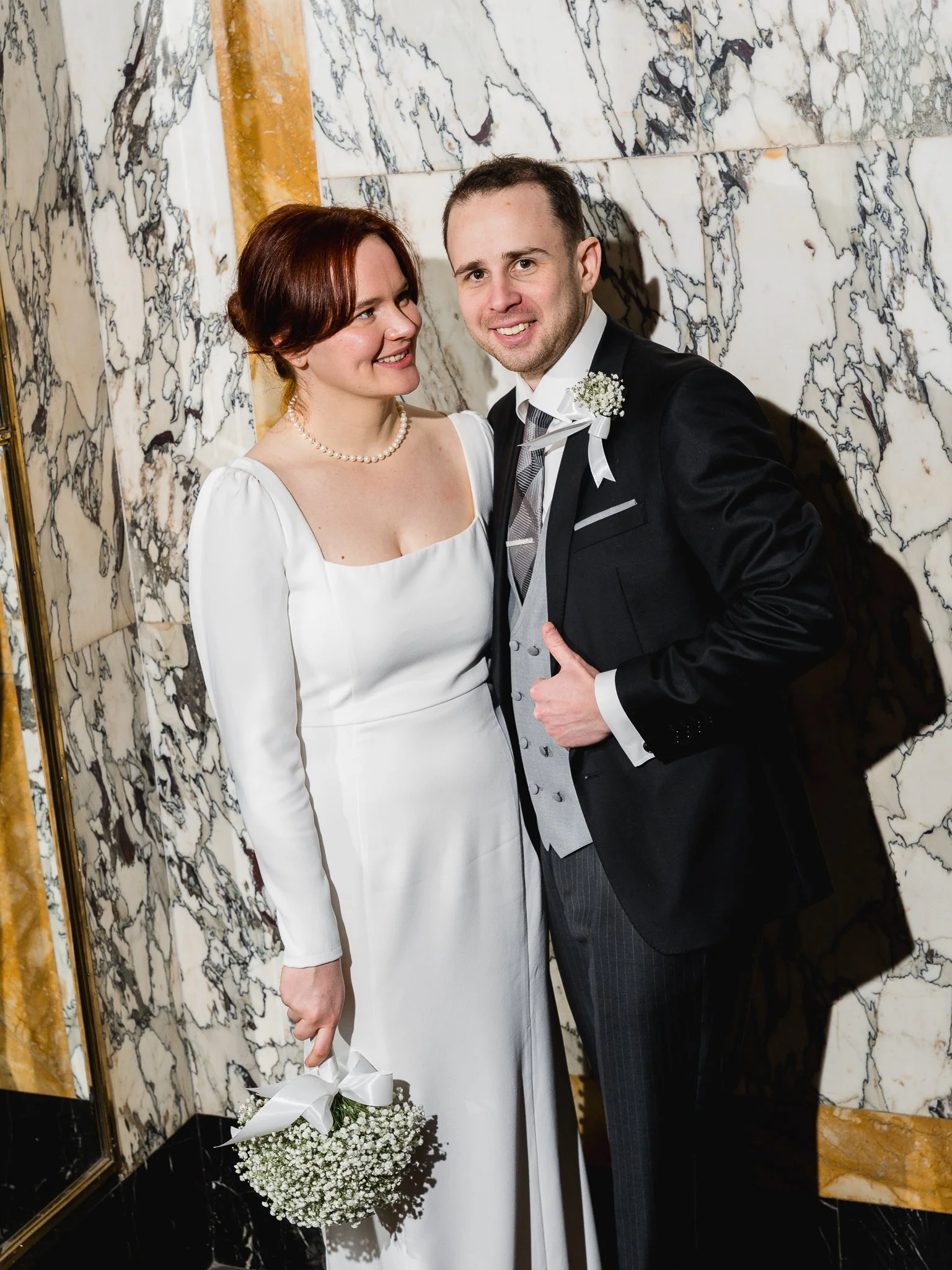 A bride and groom in wedding attire standing together against a marble wall. The bride is holding a bouquet of baby's breath with a white ribbon, and the groom is giving a thumbs-up.