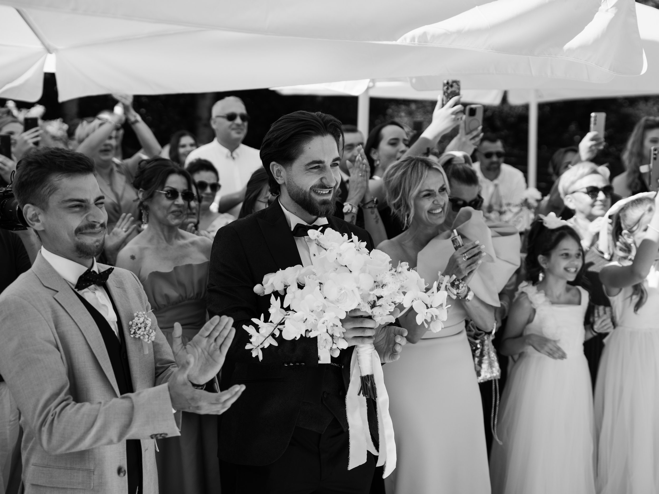 Scene from a wedding ceremony outdoors, including the groom with a beard and tuxedo, surrounded by guests clapping and taking photos, under large umbrellas.