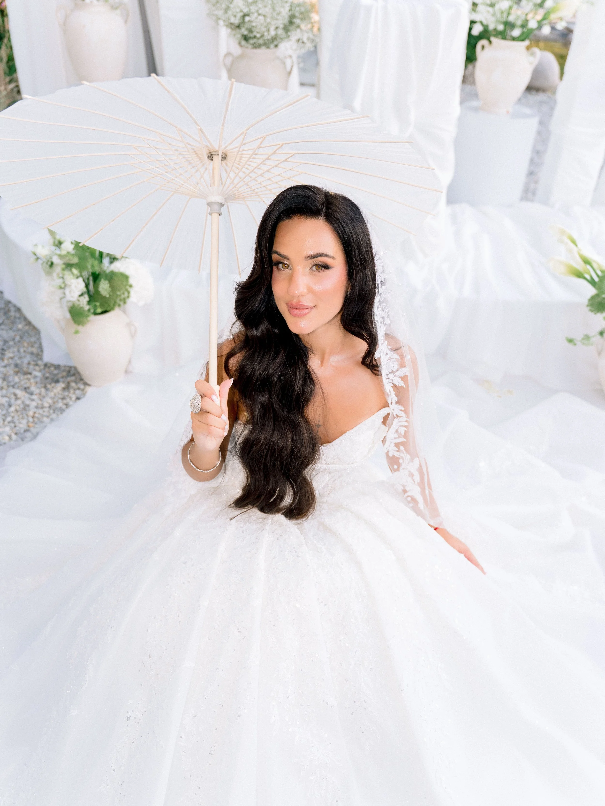 A woman dressed in a wedding gown sitting on a white surface, holding a white parasol, surrounded by white flowers and vases in a bright, decorated setting. Editorial pose taken at Croatian wedding by In Principio Weddings photography studio.