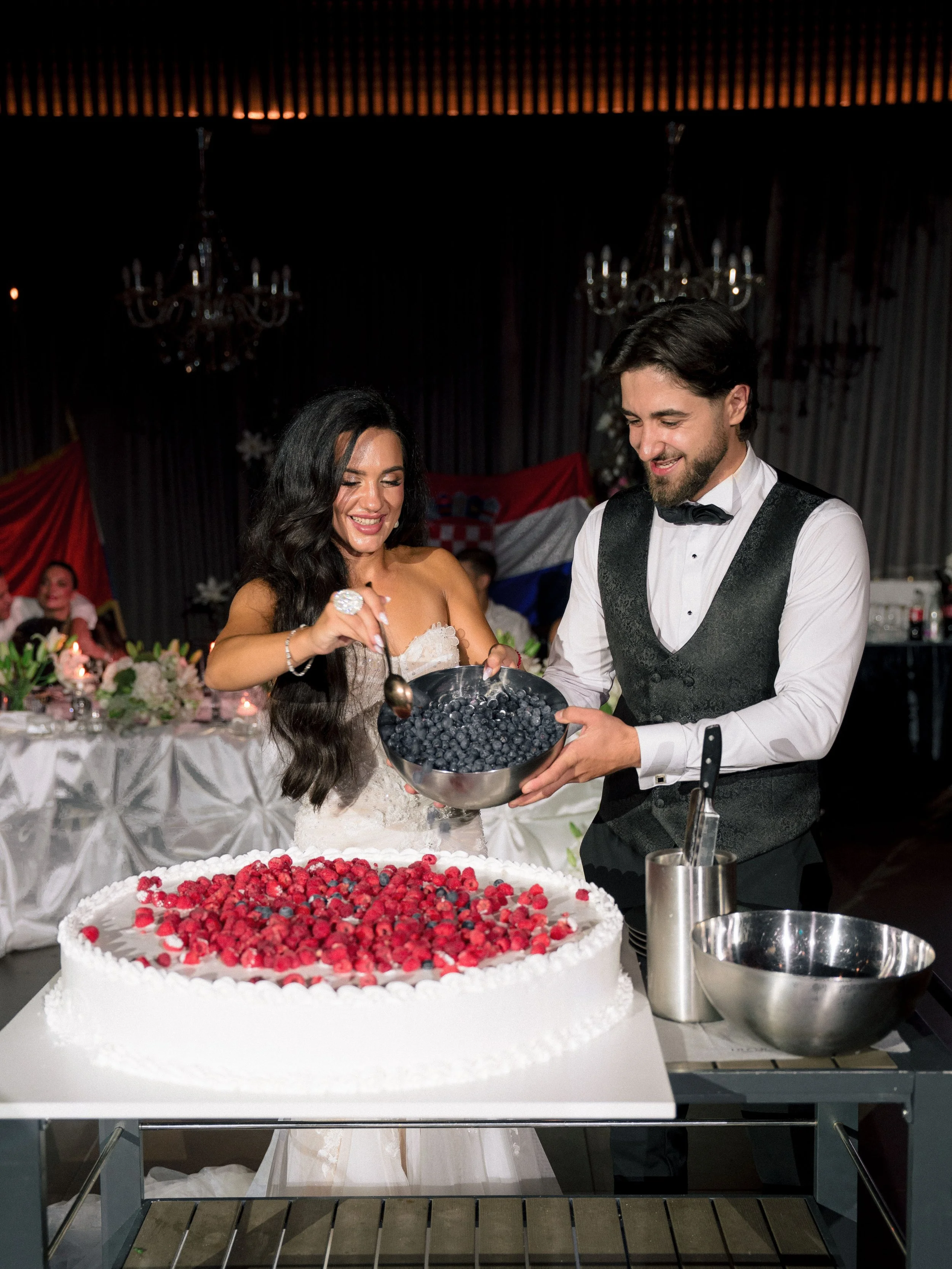 A bride and groom participate in a wedding cake cutting ceremony with a large cake topped with red and blue berries in front of them. The bride wears a white wedding dress and the groom wears a black vest and bow tie. The setting includes decorated t