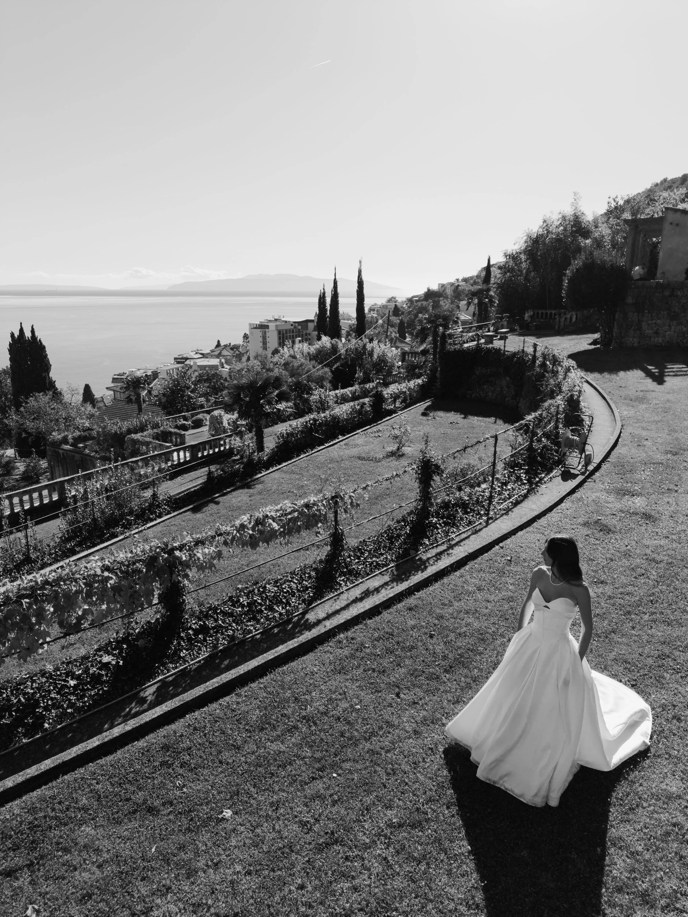 A woman in a white wedding gown standing on a grassy area with a view of a hillside and water in the background, with trees and structures around.
