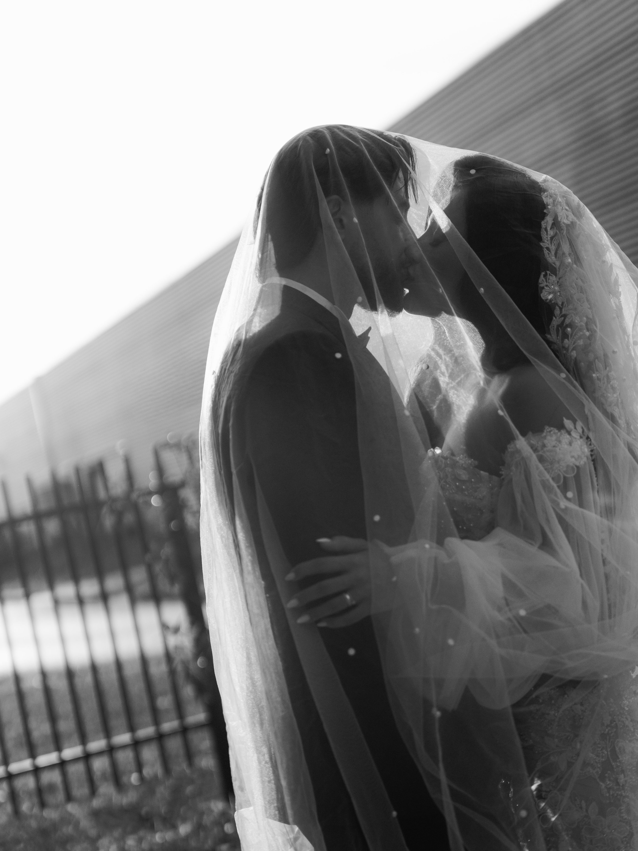 Black and white photo of a bride and groom sharing a kiss, with the bride wearing a veil and lace wedding dress, and the groom in a tuxedo, outdoors near a fence and a building.