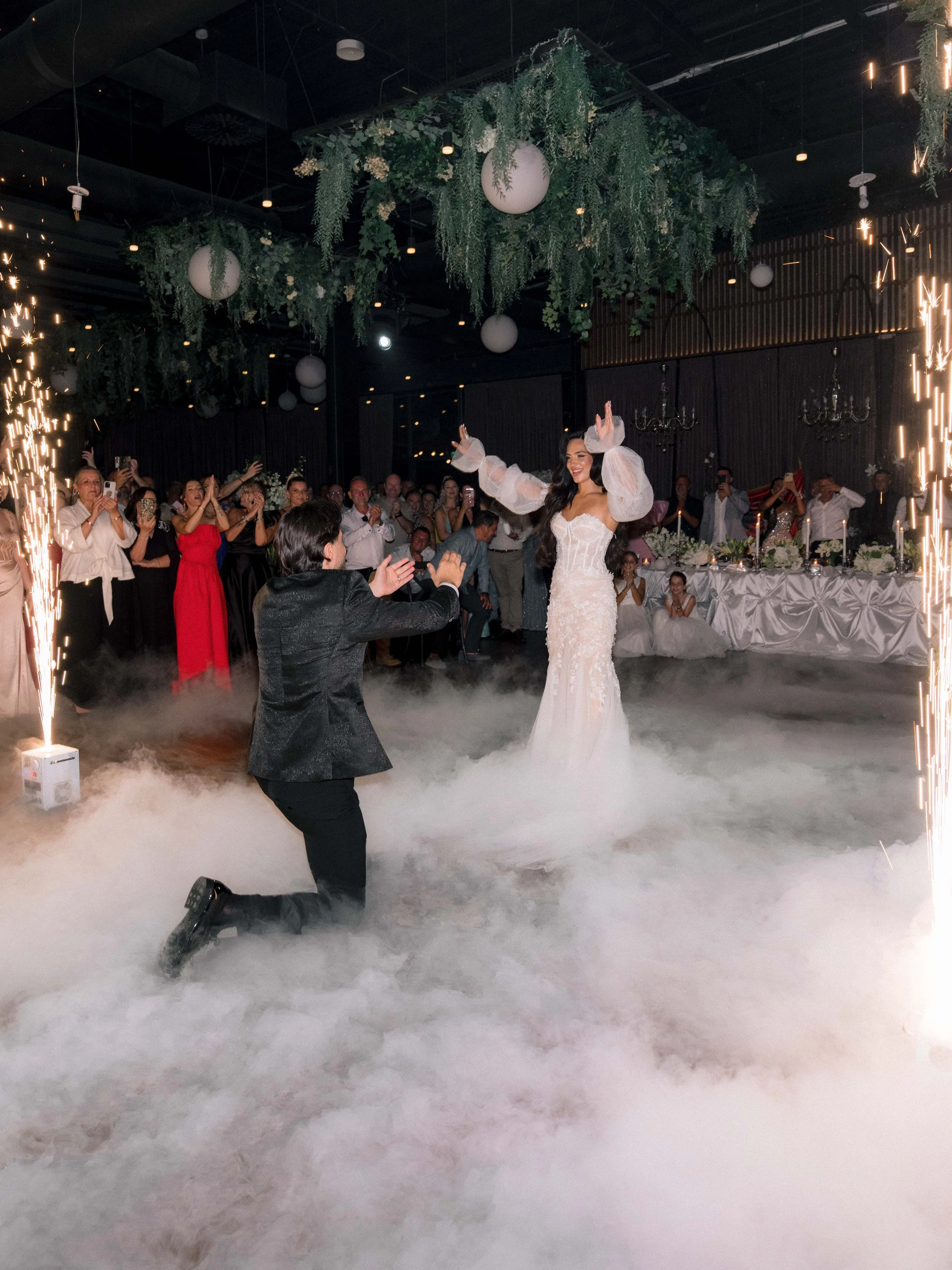 A bride and groom celebrate with guests at a wedding reception, with the groom kneeling and holding the bride's hand as she stands and smiles. There are sparklers and fog on the dance floor, with guests watching in the background.